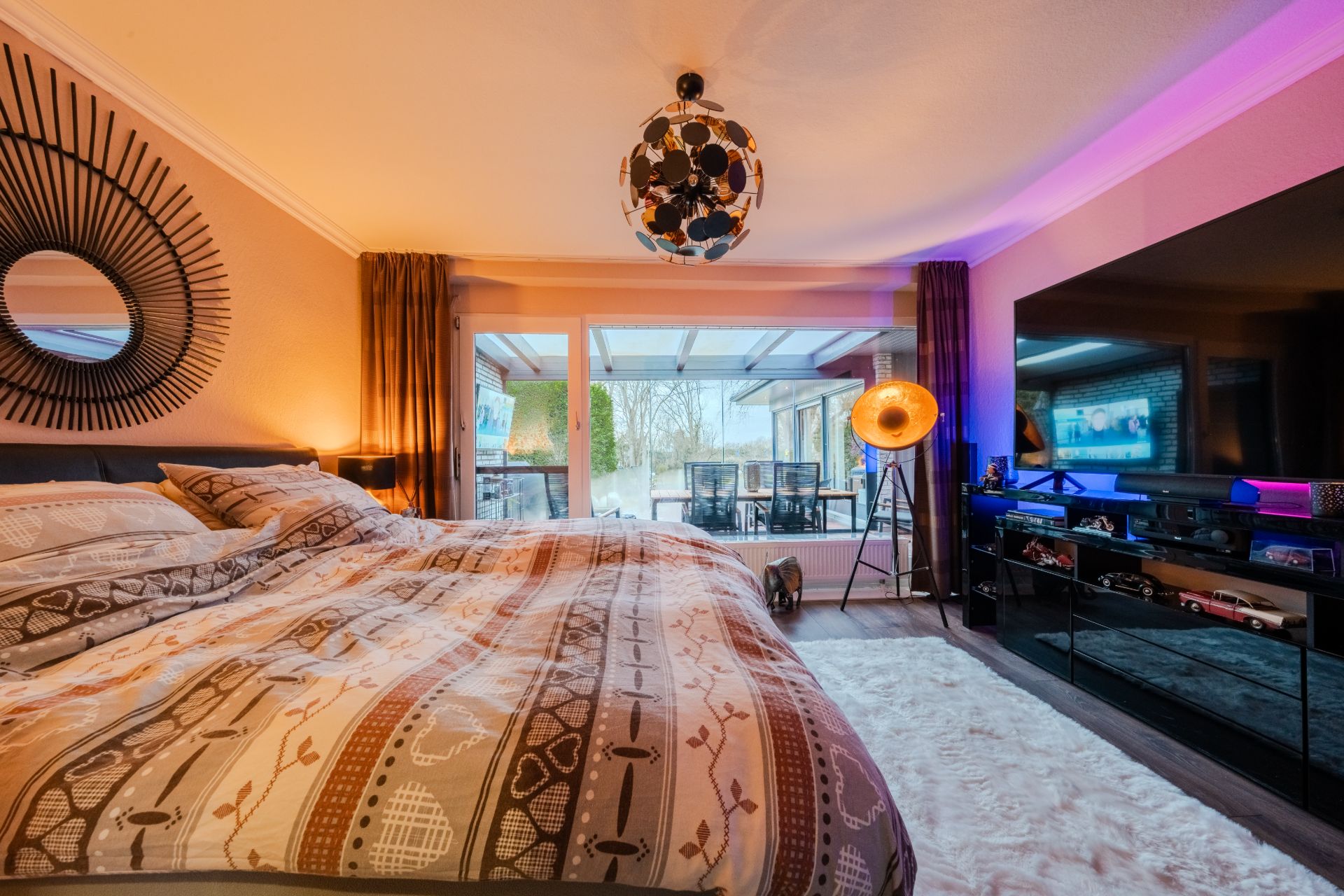 Modernes Schlafzimmer mit großem Bett, Spiegel, Fernseher und Blick auf eine Terrasse.