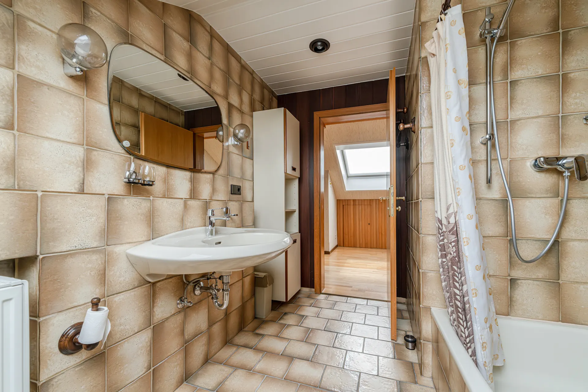 Bad mit beigem Fliesenspiegel, Waschbecken, Spiegel, Badewanne mit Duschvorhang und offenem Türdurchgang.
