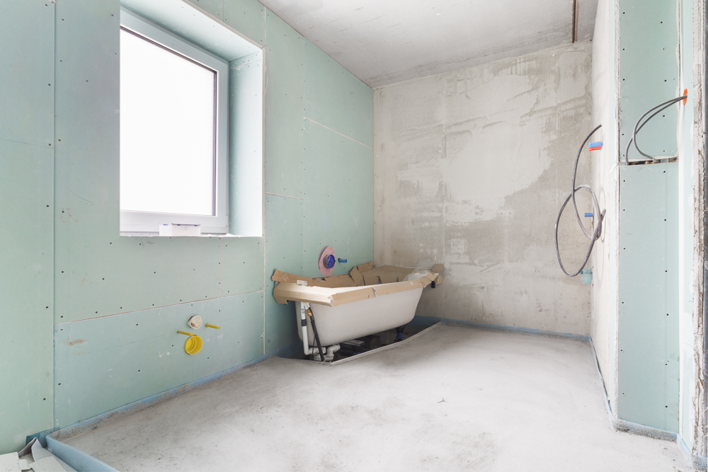 Unfertiges Badezimmer mit Badewanne, Fenster und Rohren an den Wänden.