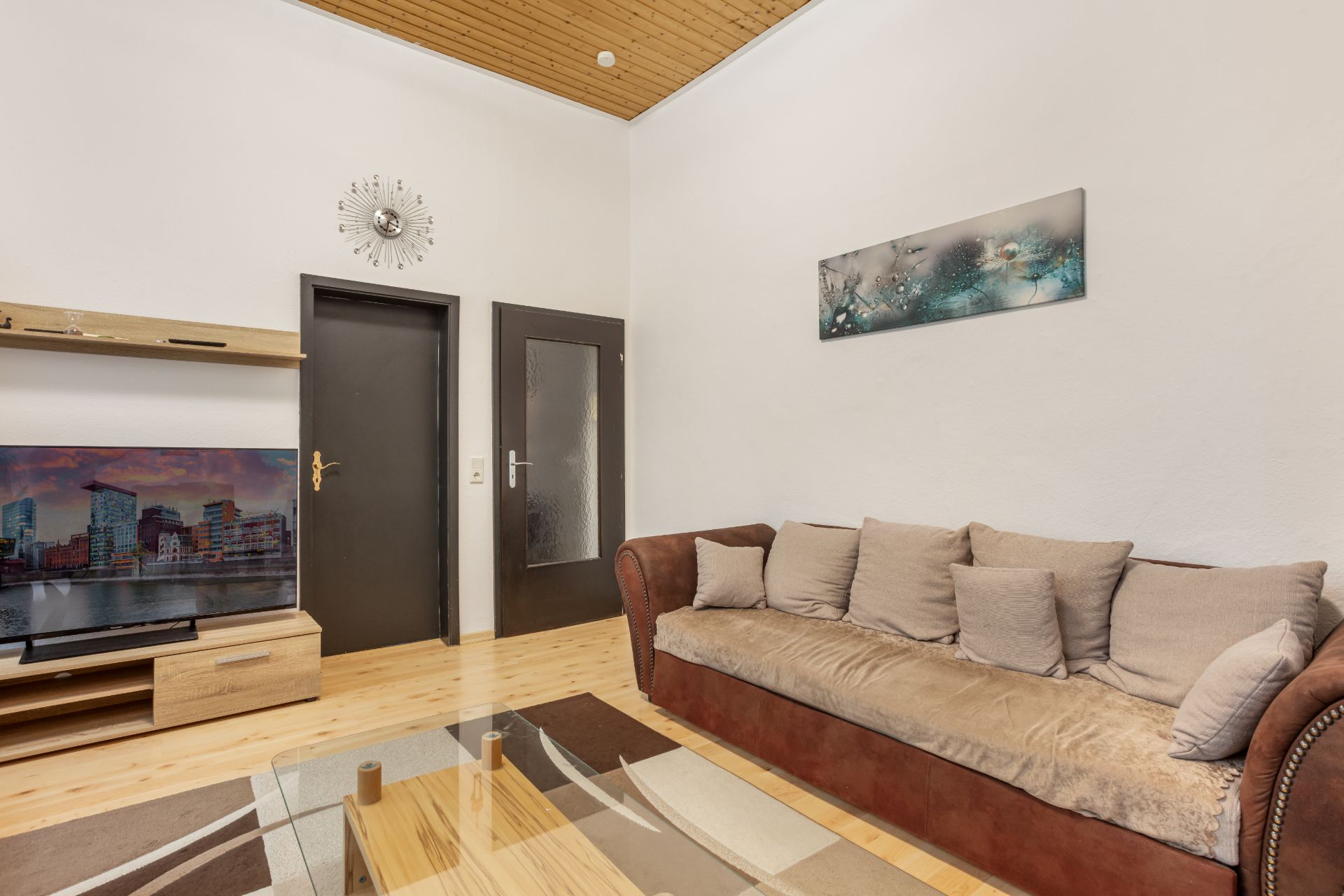 Wohnzimmer mit braunem Sofa, Glastisch, TV, zwei Türen und Wandbild.