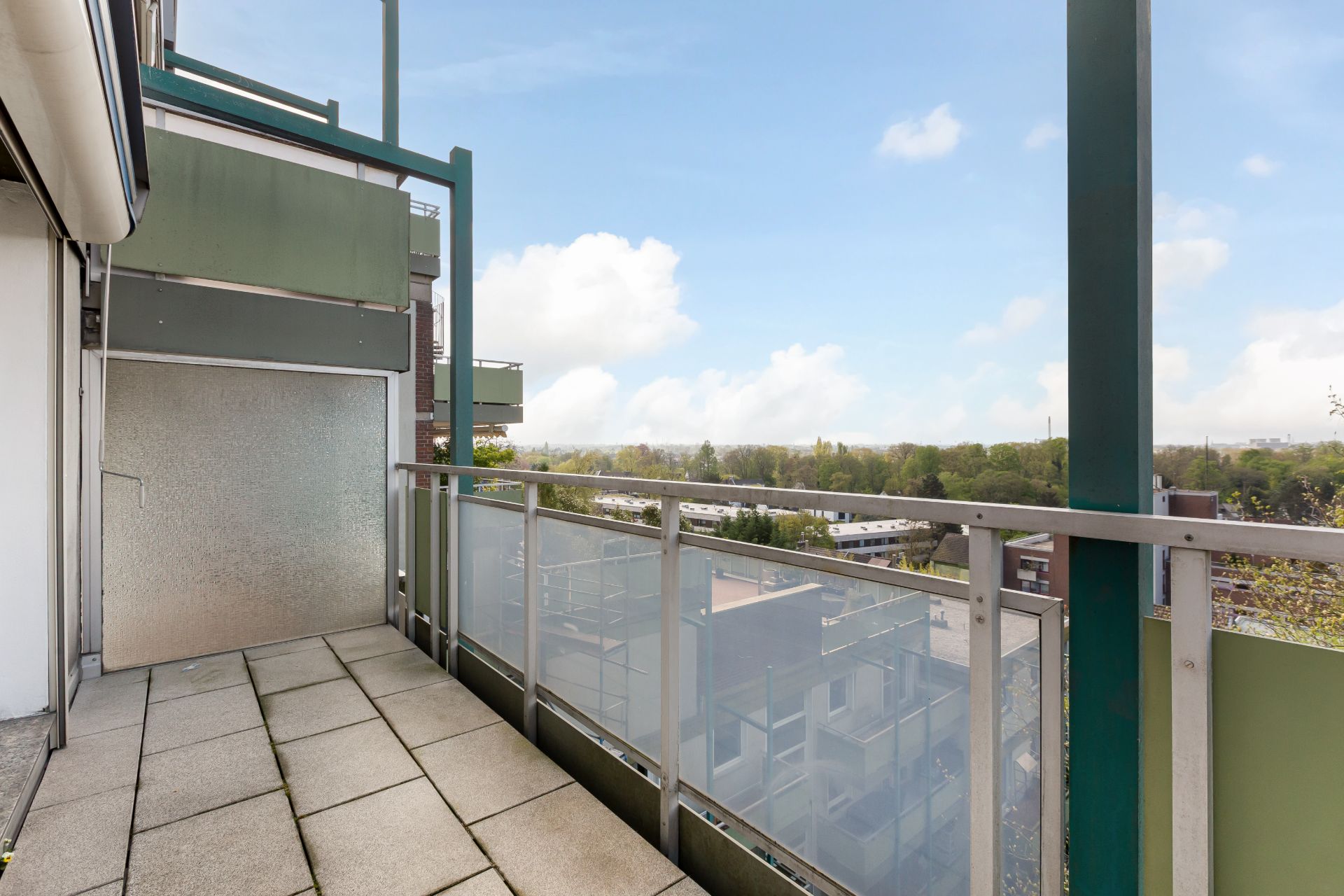 Balkon mit Glasgeländer und Blick auf grüne Bäume unter blauem Himmel.