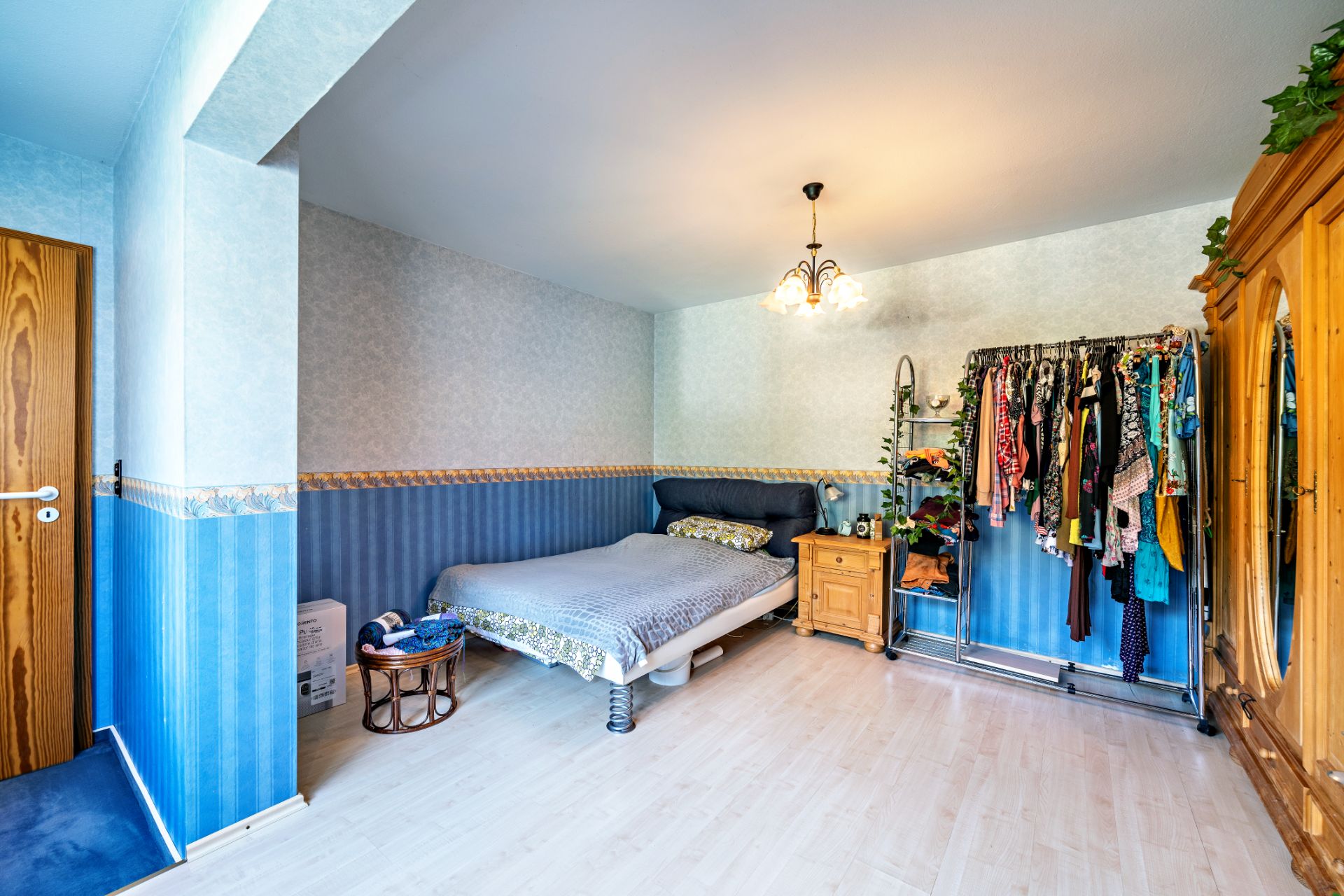 Schlafzimmer mit Doppelbett, Kleiderständer, Holzschrank und Nachttisch, blau tapezierte Wände.
