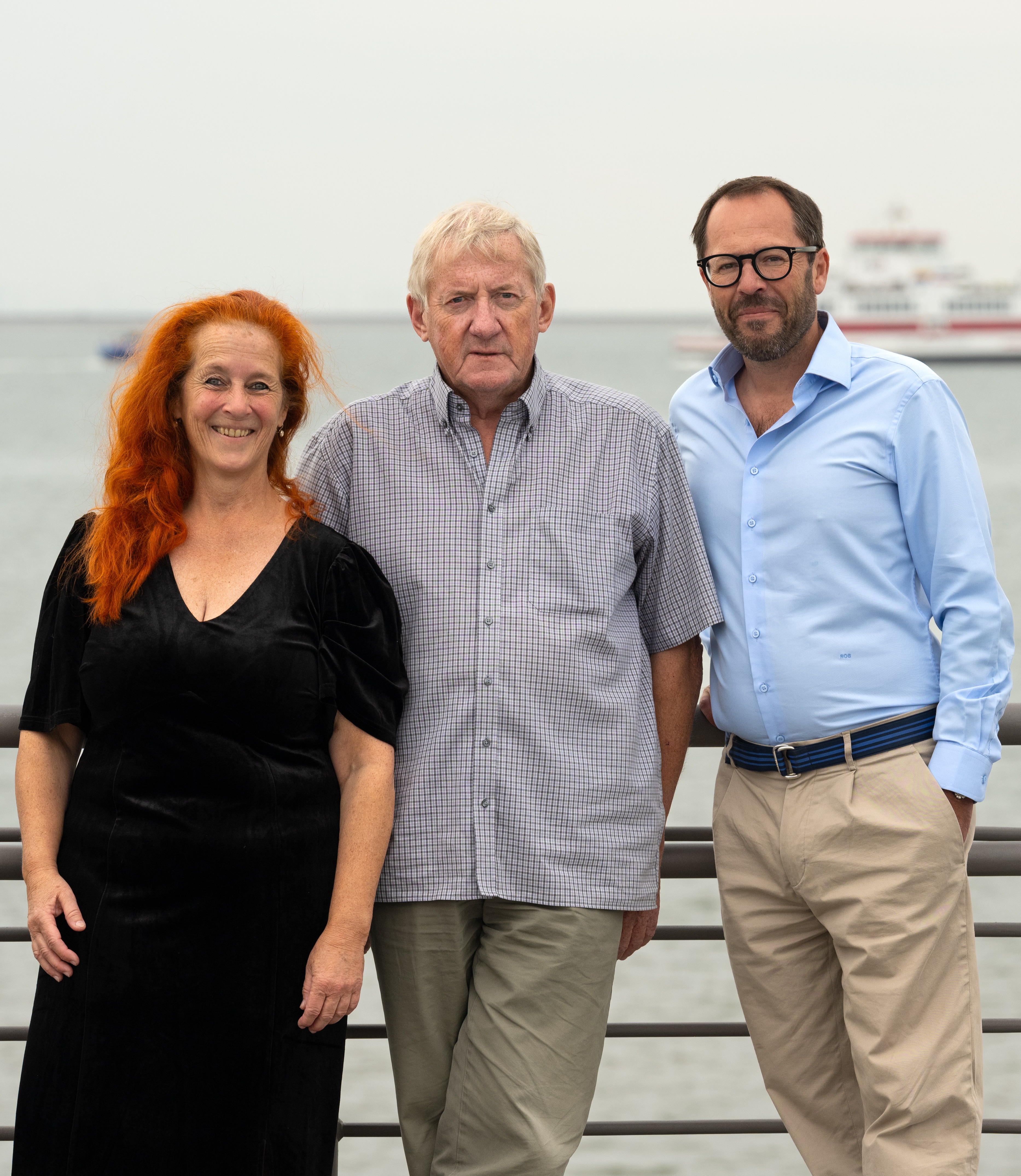 Drei Erwachsene stehen an einem Geländer mit Wasser und einem Schiff im Hintergrund.