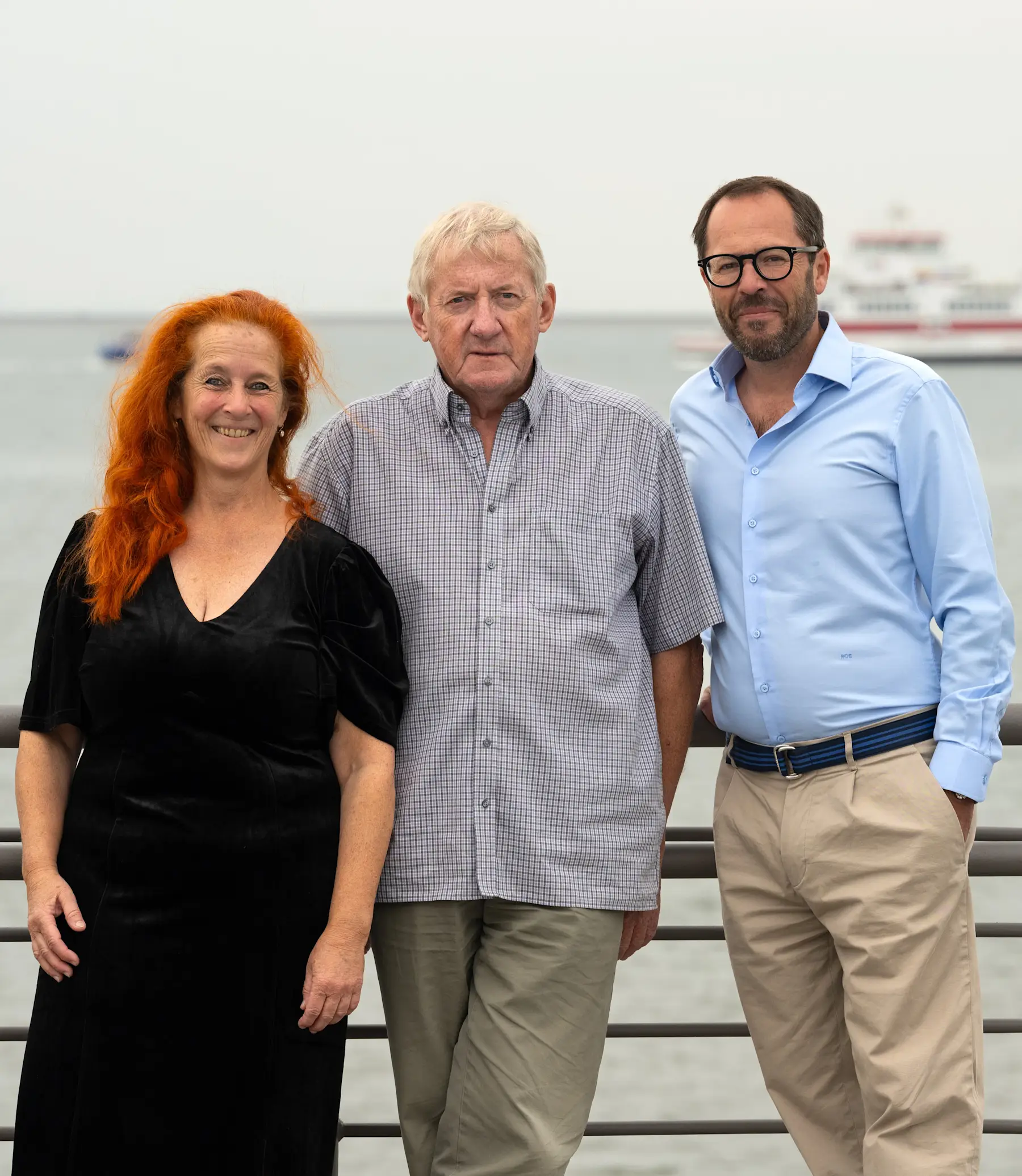 Drei Erwachsene stehen an einem Geländer mit Wasser und einem Schiff im Hintergrund.