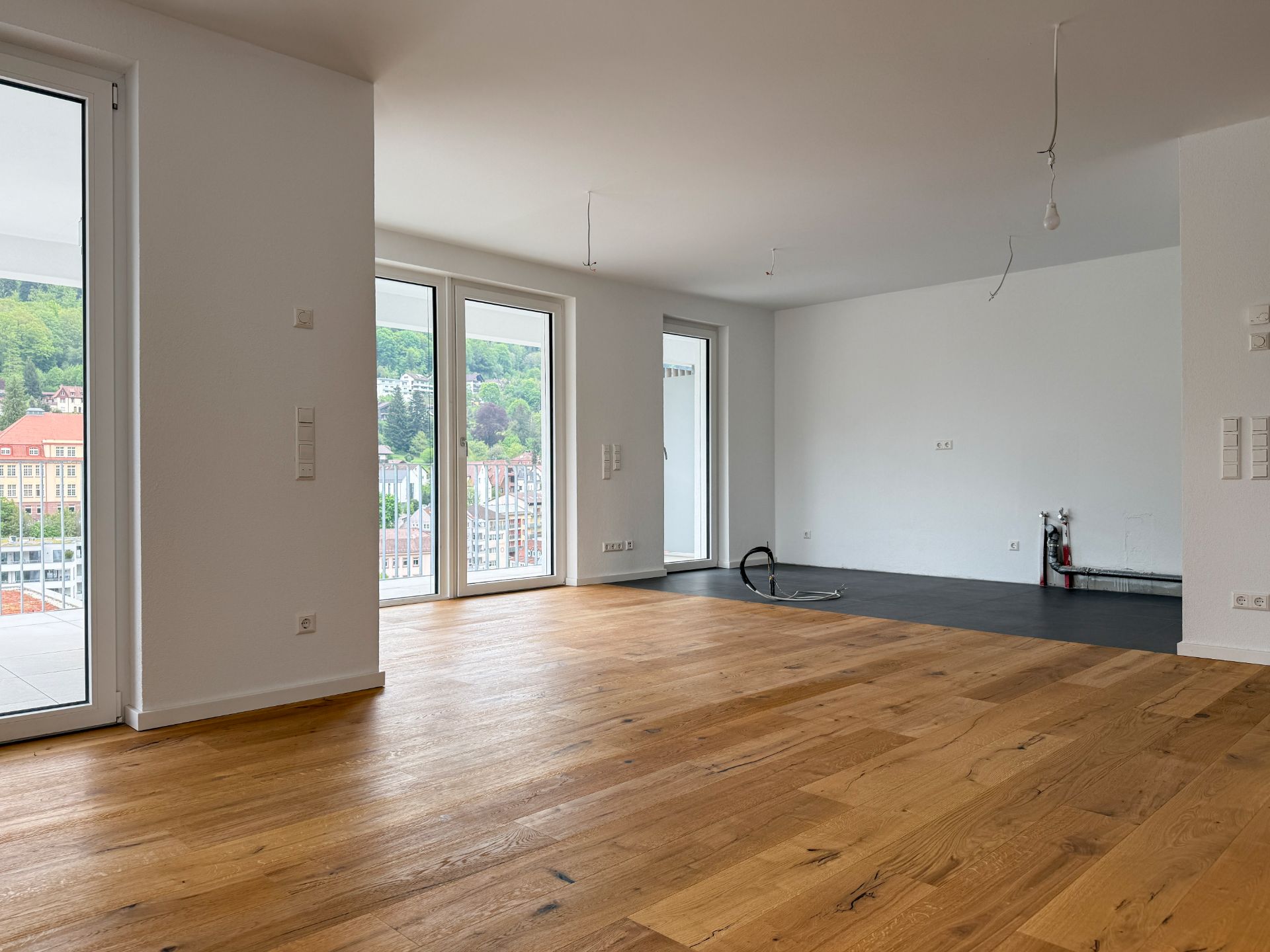 Helles, leeres Wohnzimmer mit Parkettboden und großen Fenstern, Blick auf Balkon und grüne Landschaft.