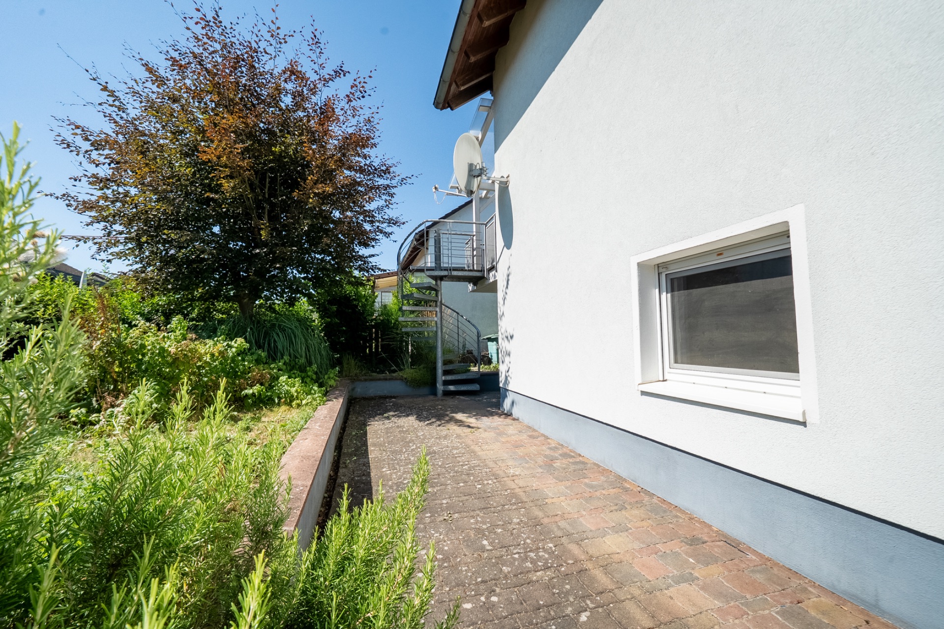 Gepflasterter Weg neben einem Wohnhaus, mit Pflanzen und einer Wendeltreppe im Hintergrund.