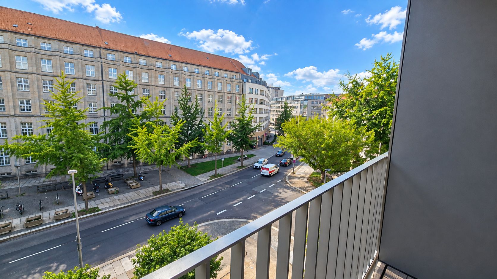 Blick vom Balkon auf eine Straße mit Autos, Bäumen und einem großen, alten Gebäude gegenüber.