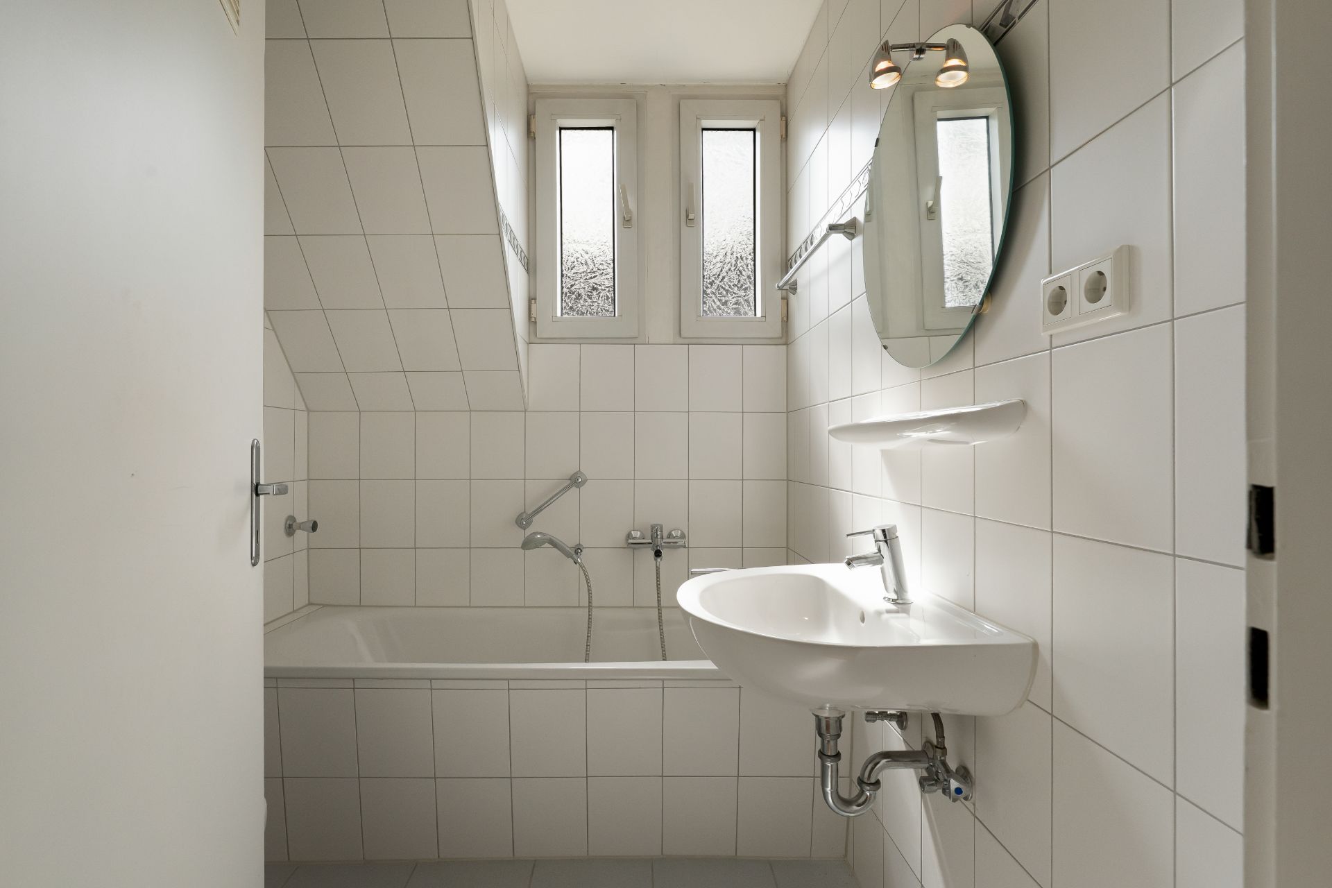 Helles Badezimmer mit weißen Fliesen, Badewanne, Waschbecken und Spiegel unter zwei Fenstern.
