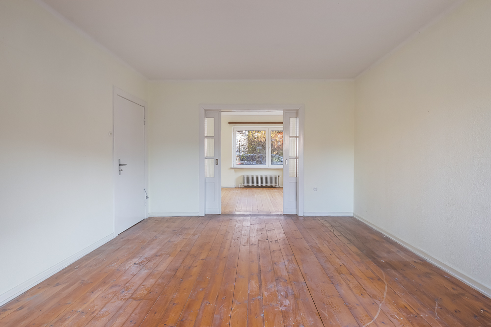 Leerer Raum mit Holzboden, weißen Wänden und Blick ins nächste Zimmer mit großem Fenster.