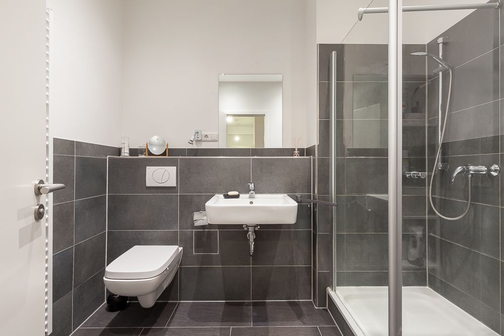 Modernes Bad mit Hänge-WC, Waschbecken und Glasdusche, graue Fliesen und weißer Wand.