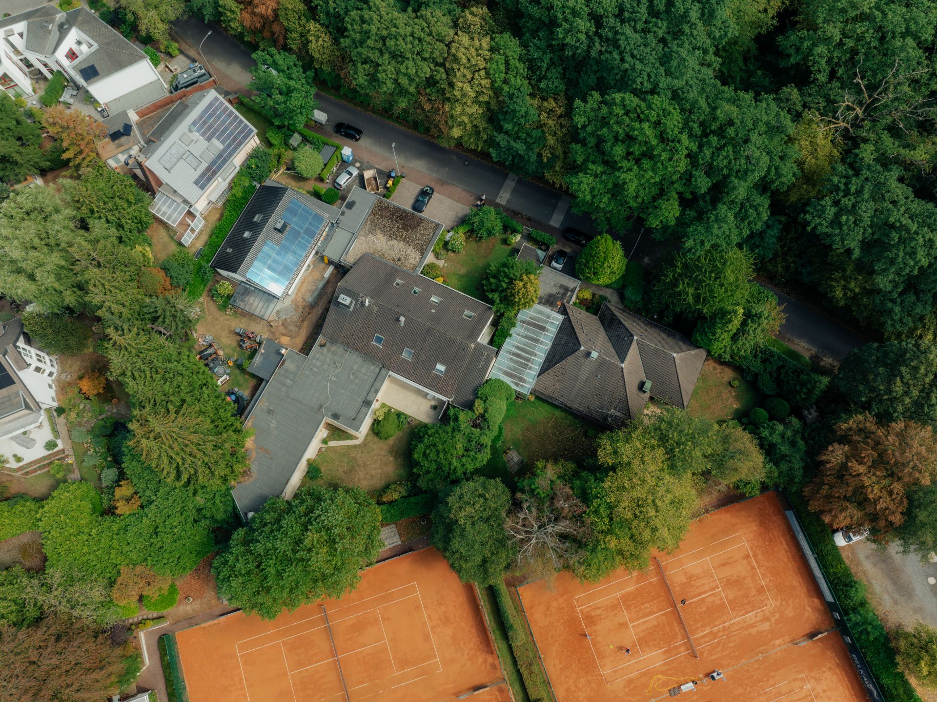 Luftaufnahme von Wohnhäusern, Garten, Straße und zwei Tennisplätzen, umgeben von Bäumen.
