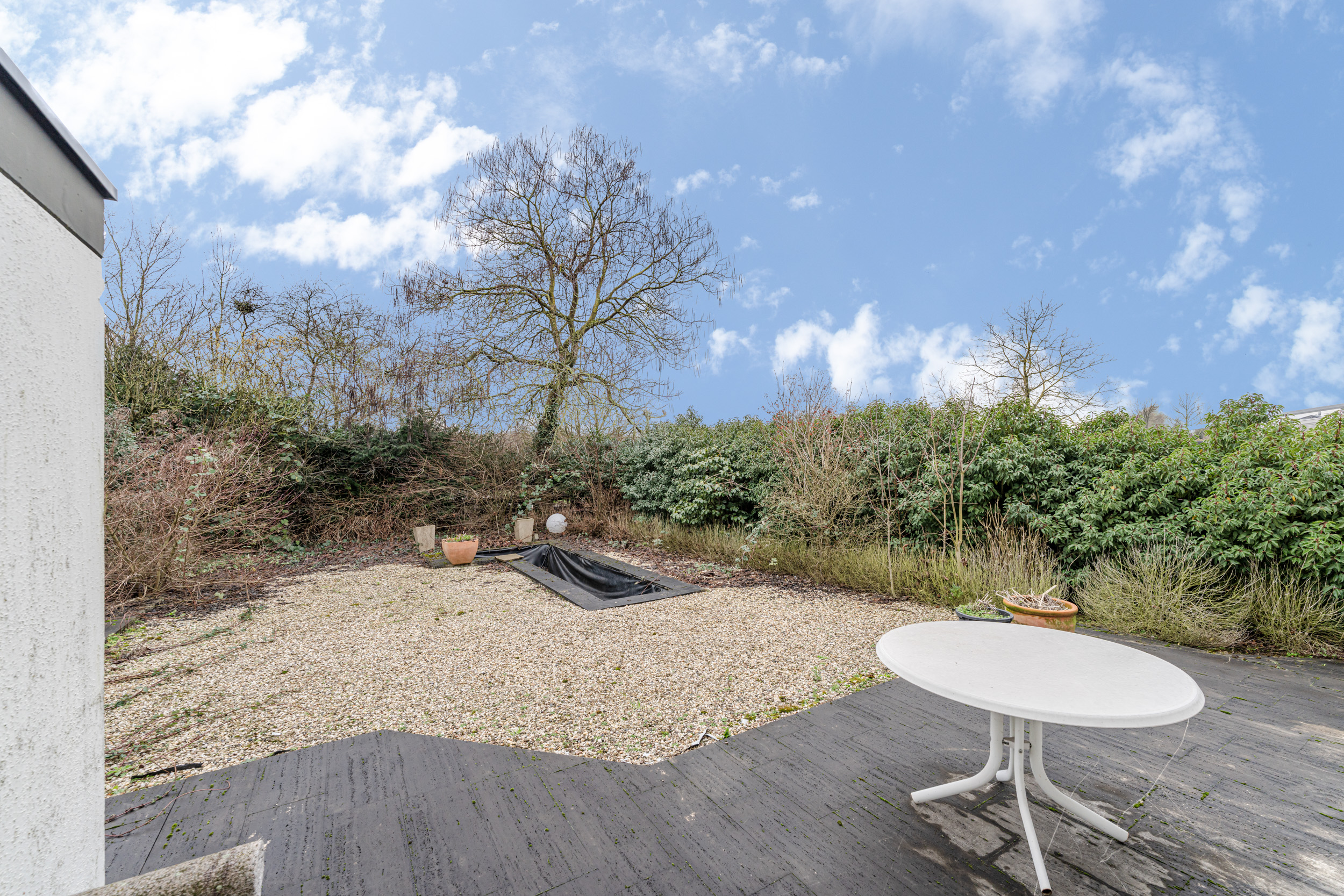 Garten mit Kiesfläche, kleinem Teich, Büschen und weißem Gartentisch auf Terrasse.