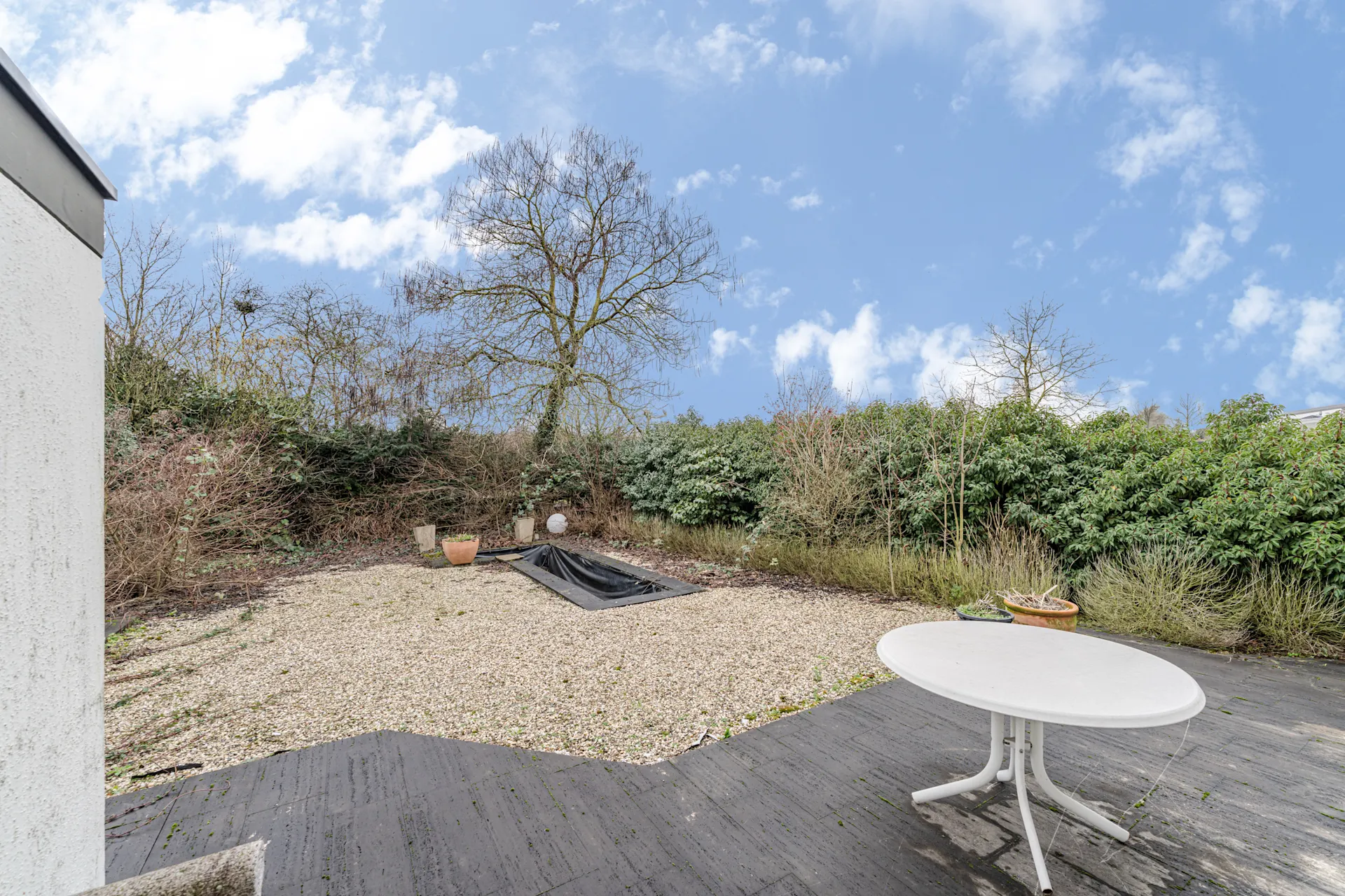 Garten mit Kiesfläche, kleinem Teich, Büschen und weißem Gartentisch auf Terrasse.