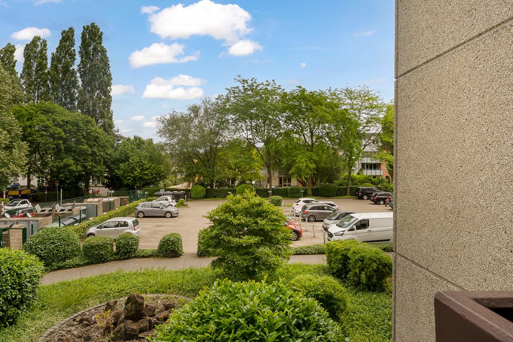 Blick auf einen Parkplatz mit mehreren Autos und umgebenden Bäumen, aufgenommen von einem Balkon.