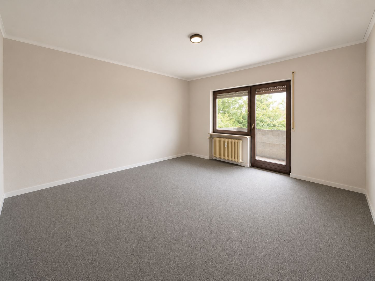 Leeres Zimmer mit grauem Teppich, großem Fenster und Tür zum Balkon, darunter ein Heizkörper.