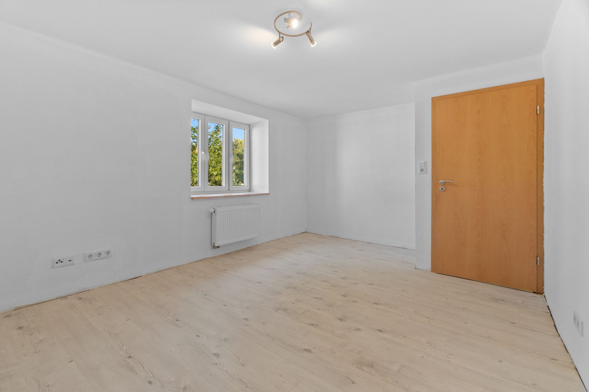 Helles, leeres Zimmer mit Holzboden, Fenster und Holztür.