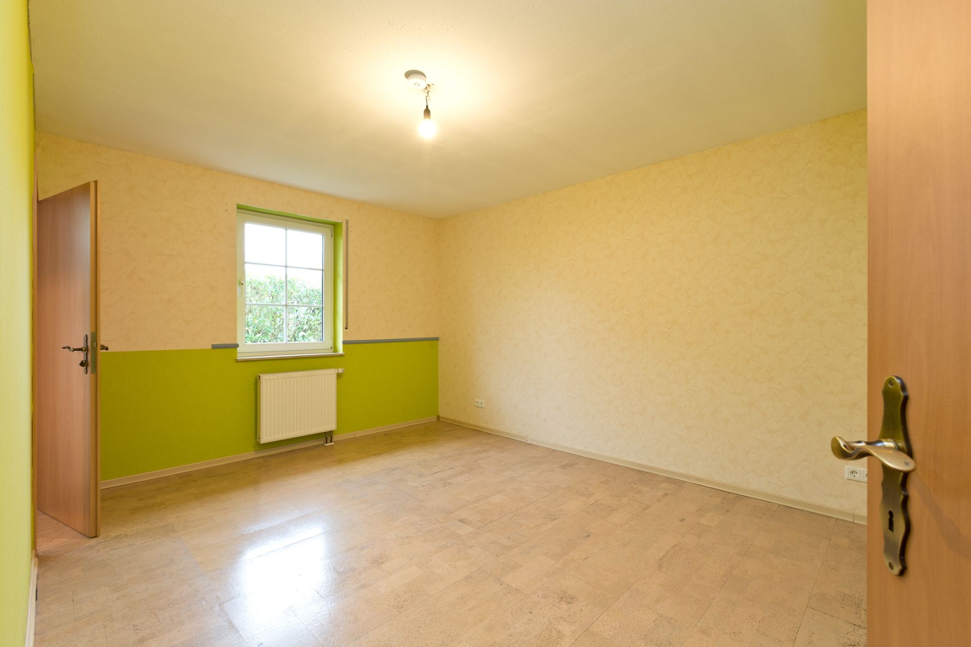 Leerer, heller Raum mit Holzfußboden, grün-gelber Wand und Fenster, Tür ist offen.
