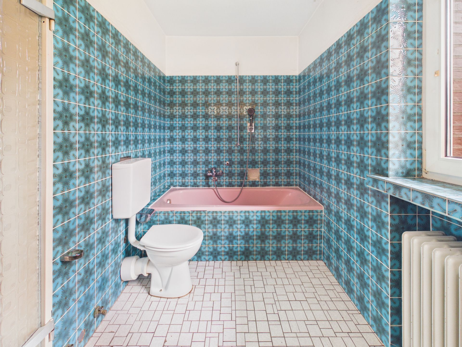 Badezimmer mit blauen Fliesen, rosa Badewanne, Toilette und Fenster.