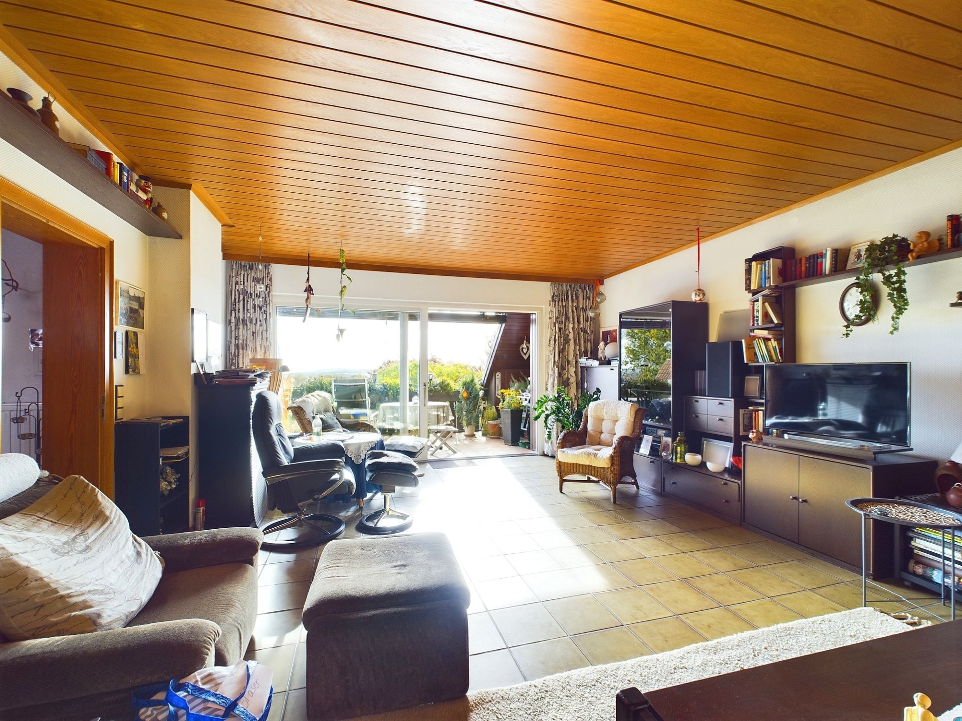 Wohnzimmer mit Holzdecke, Sofa, Sesseln, Fernseher und großen Fenstern zum Balkon.