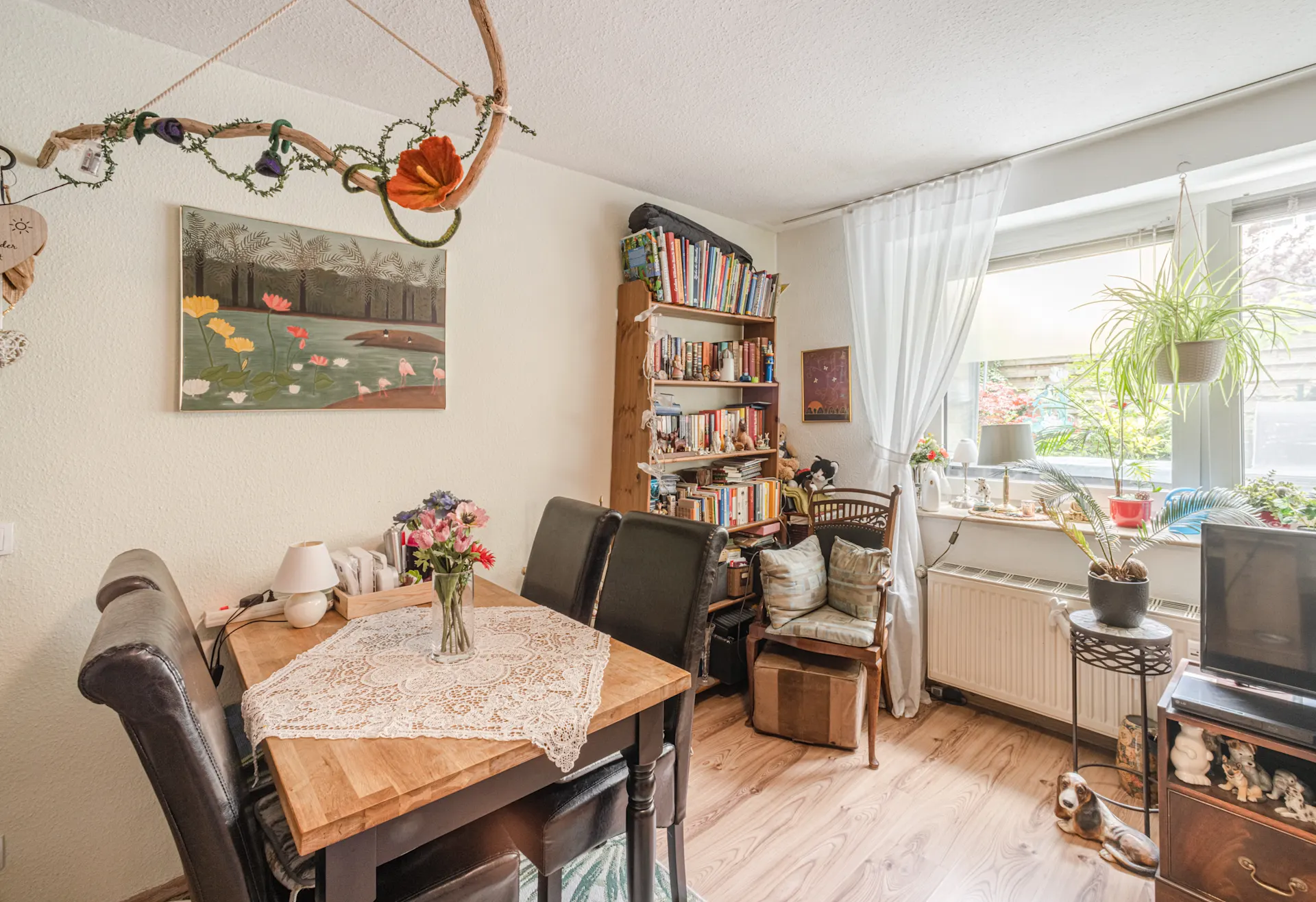 Gemütliches Wohnzimmer mit Esstisch, Bücherregal, Stühlen, Pflanzen und großem Fenster.