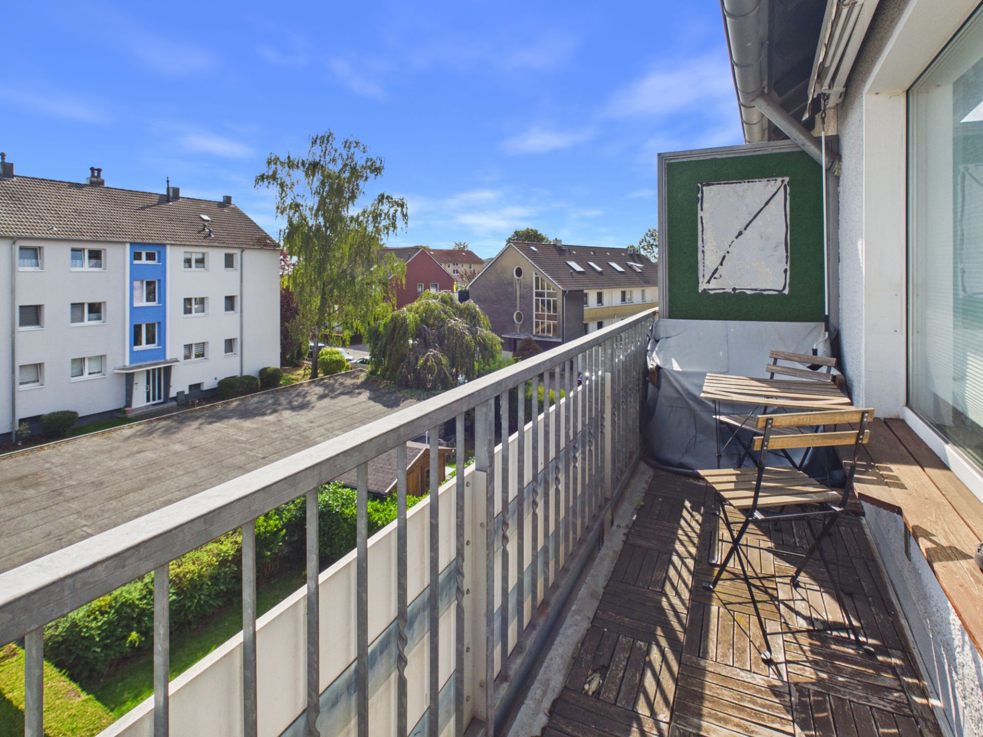 Balkon mit Holzboden, kleinem Tisch und zwei Stühlen, Blick auf Wohnhäuser und Bäume.
