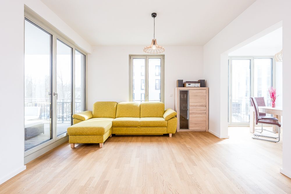 Helles Wohnzimmer mit gelbem Ecksofa, Holzboden und großem Fenster, daneben ein kleiner Holzschrank.