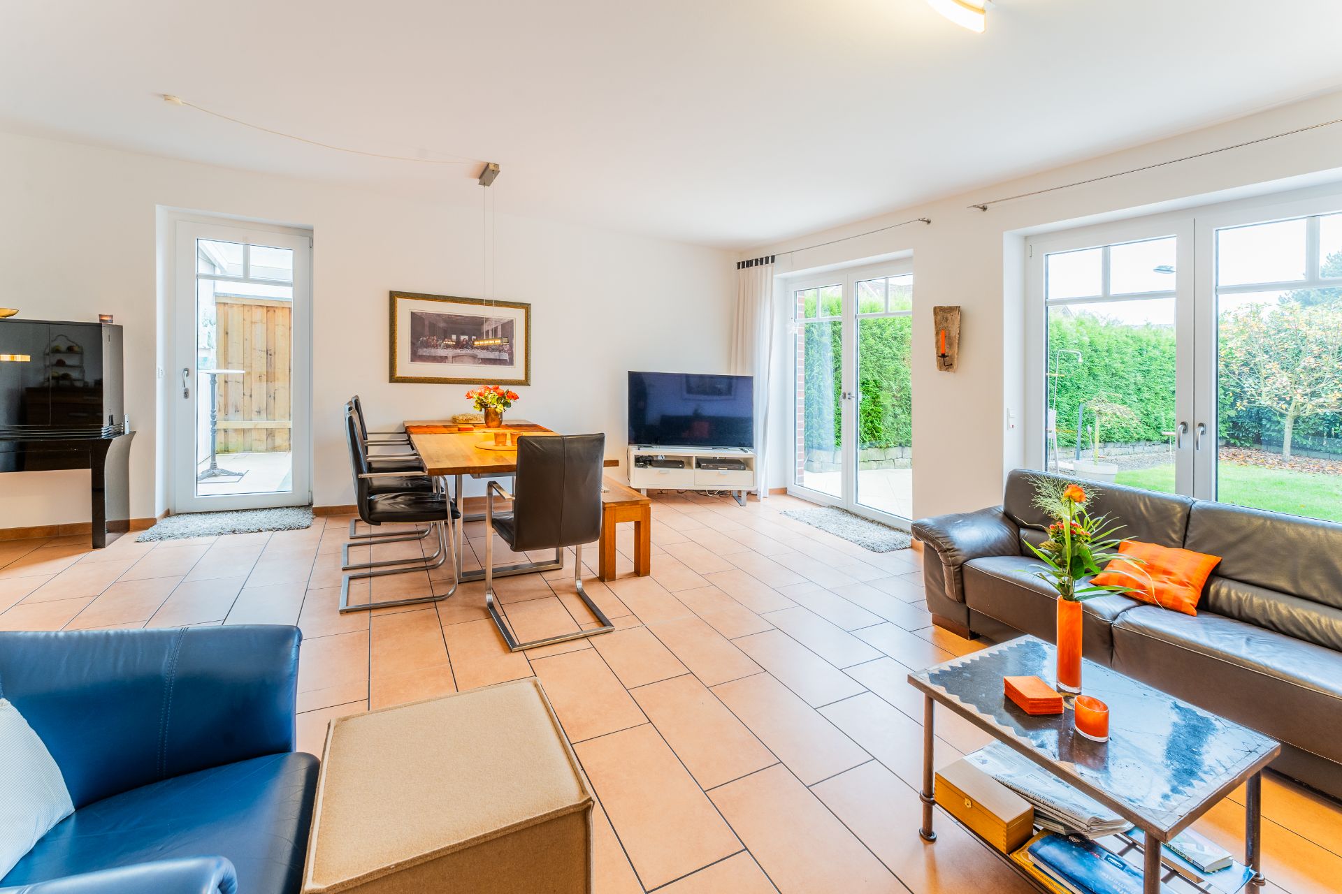 Helles Wohnzimmer mit Sofa, Esstisch, Fernseher und großen Fenstern zum Garten.