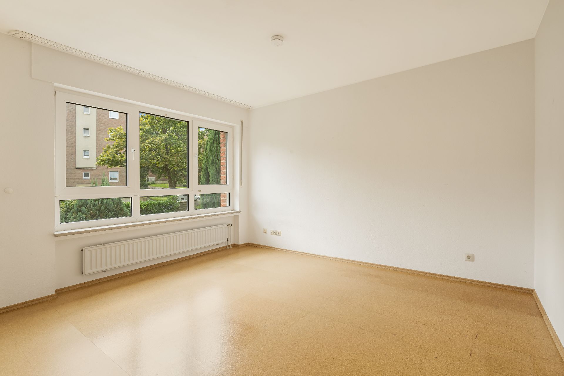 Leeres Zimmer mit großem Fenster, hellem Boden und weiß gestrichenen Wänden.