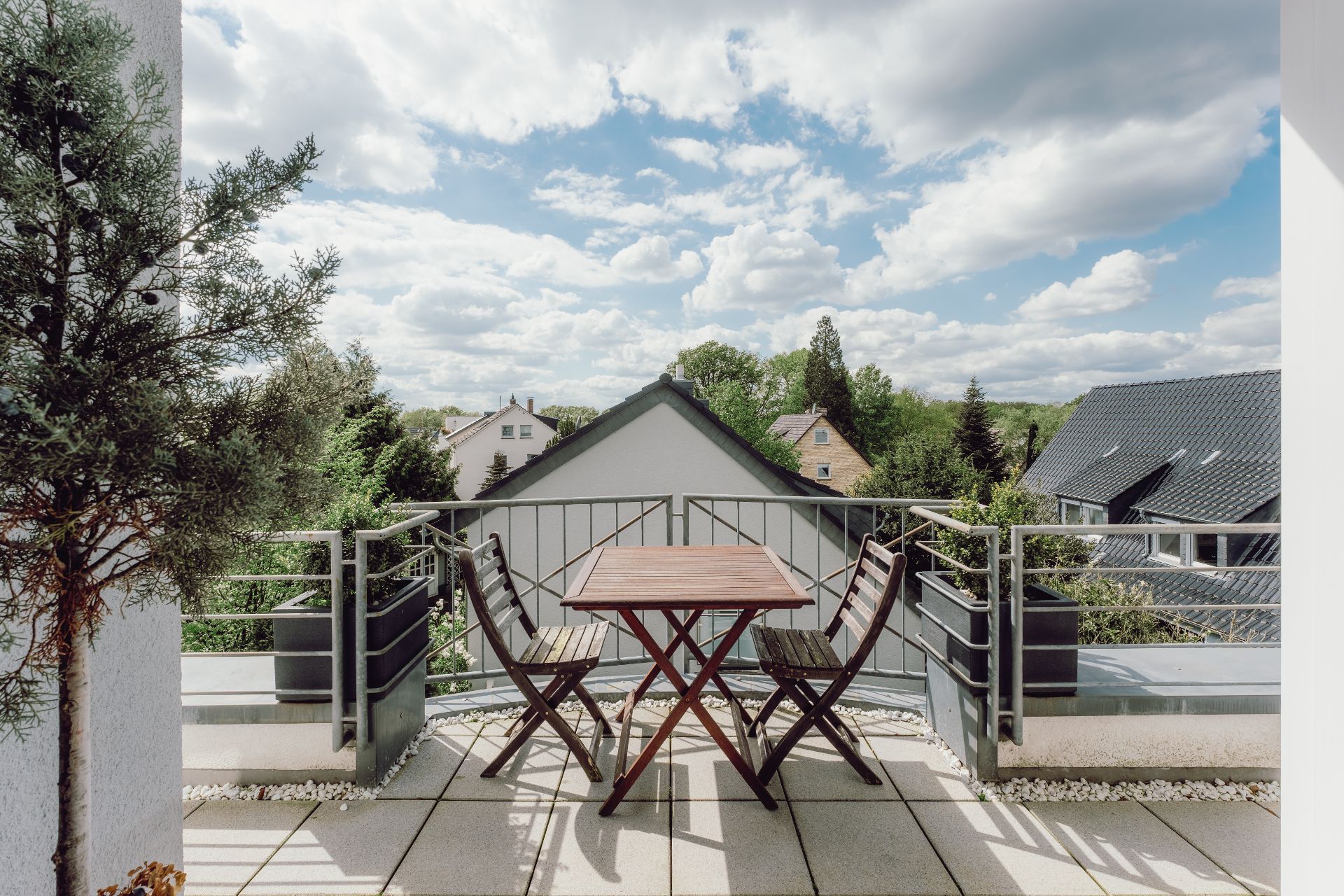 Balkon mit kleinem Holztisch und zwei Stühlen, Ausblick auf Häuser und Bäume bei Tageslicht.
