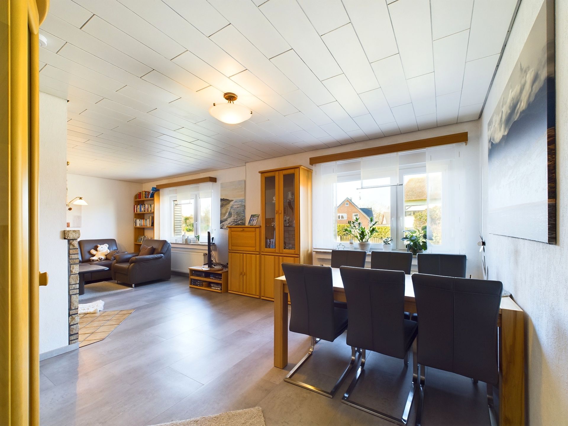 Helles Wohnzimmer mit Esstisch, sechs Stühlen, Sofaecke und großen Fenstern.