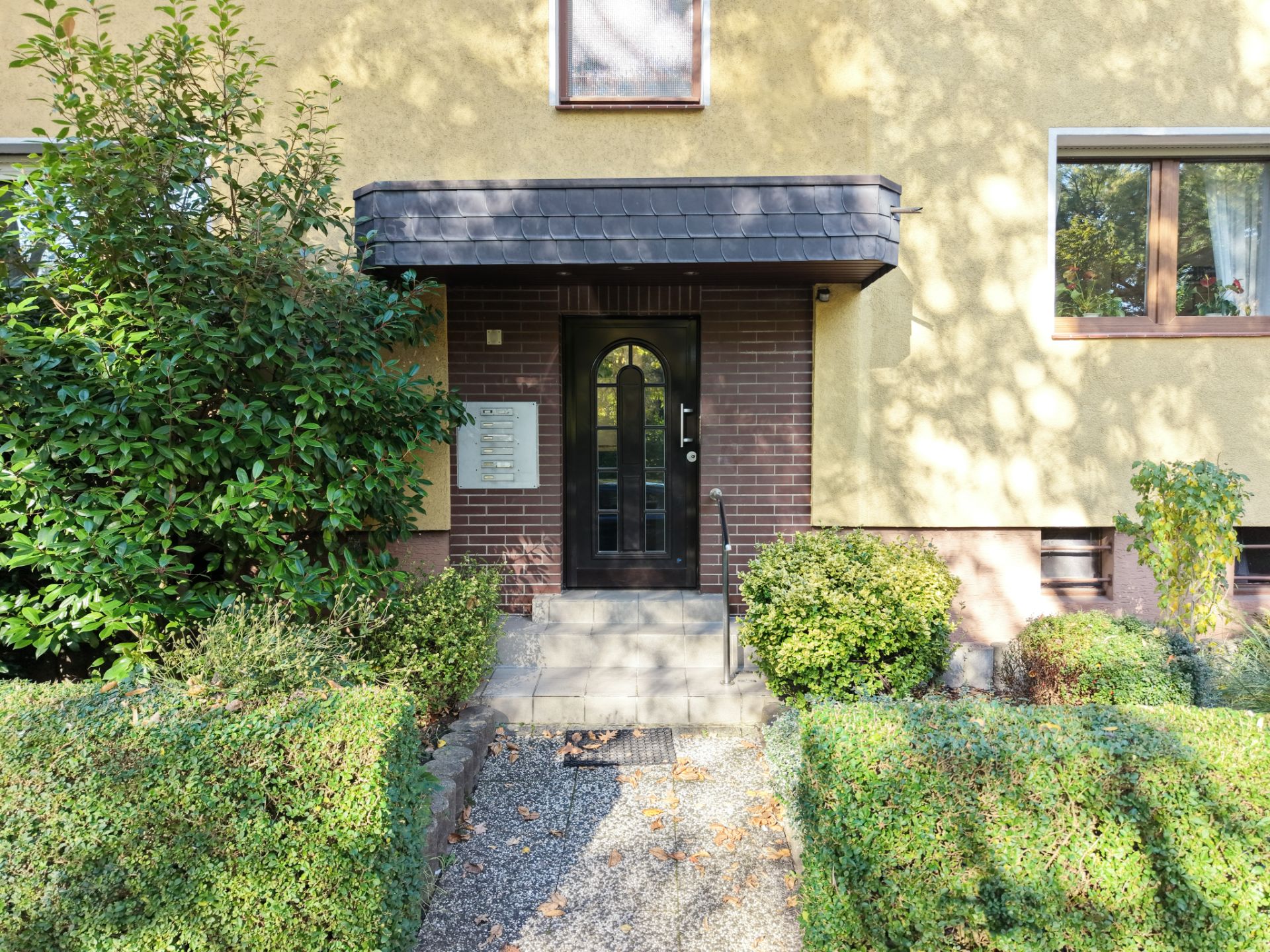 Hauseingang mit schwarzer Tür und Treppe, umgeben von Büschen und gelber Fassade.