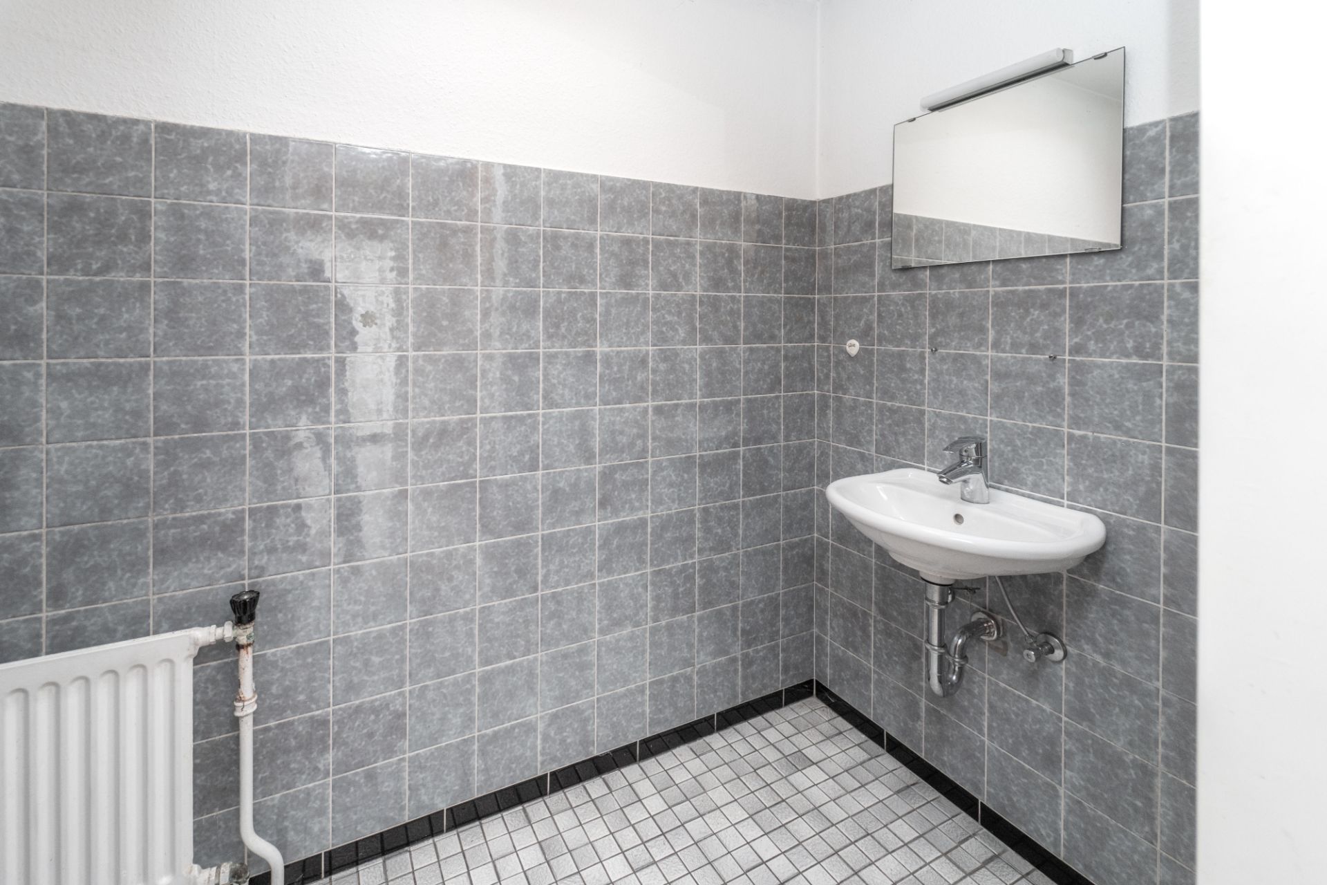 Waschbecken mit Spiegel an einer grau gefliesten Wand, Heizkörper links unten.