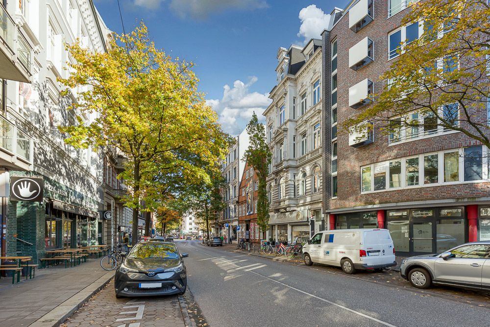 Stadtstraße mit Bäumen, geparkten Autos und Wohnhäusern an einem sonnigen Tag.