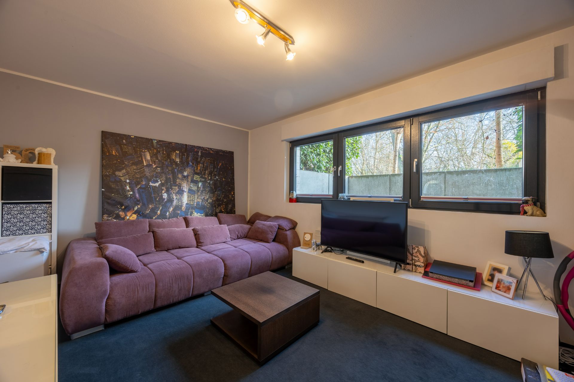 Wohnzimmer mit lila Sofa, Couchtisch, TV auf weißem Schrank und großem Fenster.
