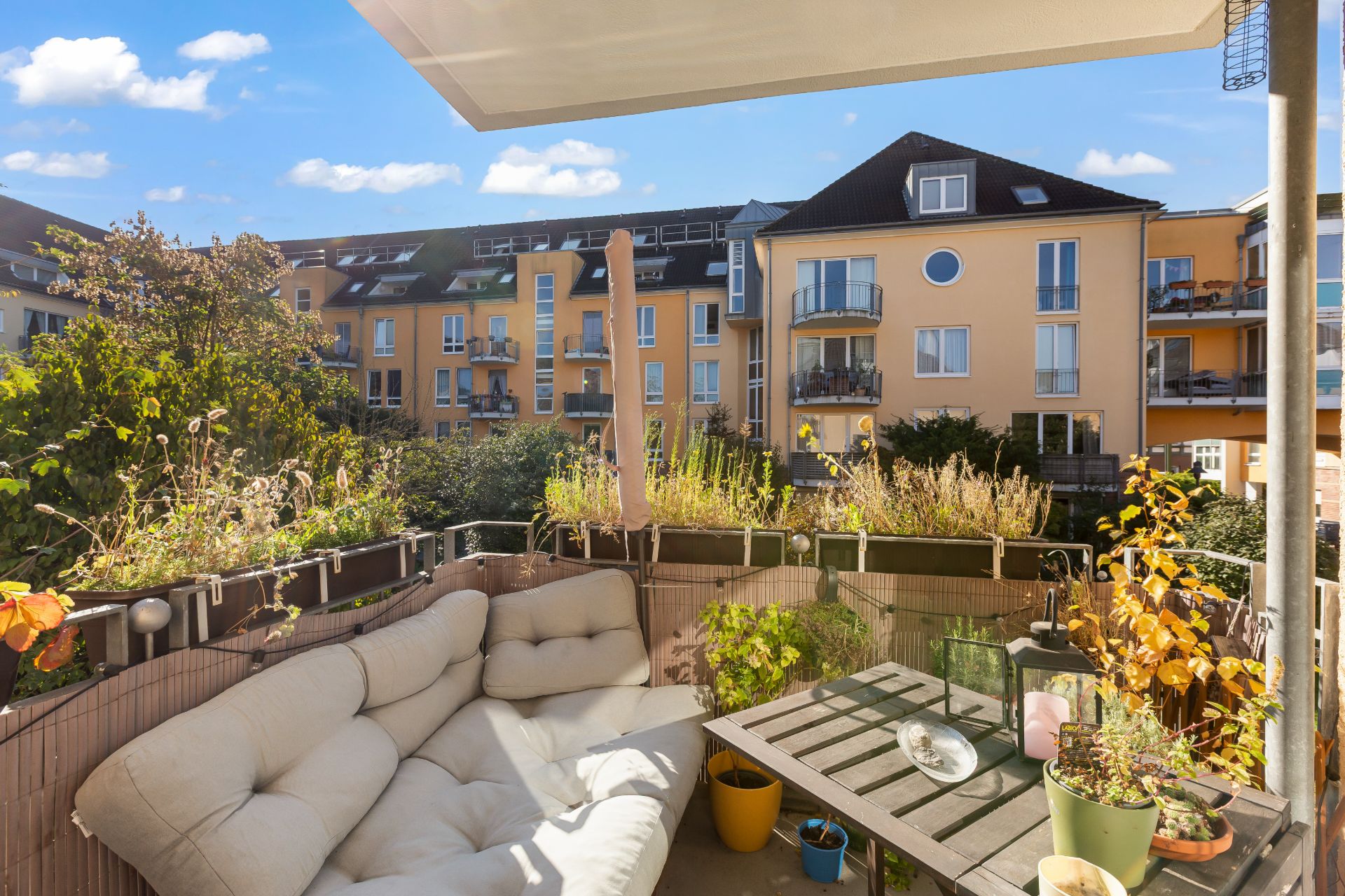 Gemütlicher Balkon mit Sofa, Tisch und Pflanzen, Blick auf gelbe Wohnhäuser im Hintergrund.