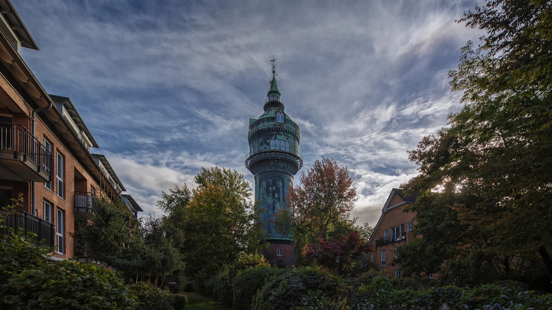 Alter Wasserturm zwischen Wohnhäusern und Bäumen unter bewölktem Himmel.