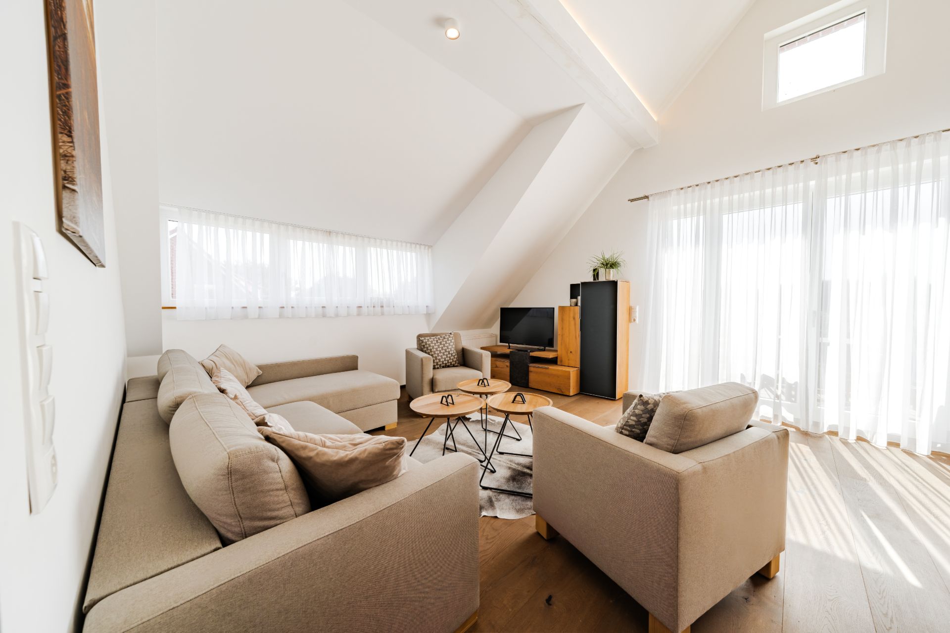Helles Wohnzimmer mit Sofa, Sesseln, Fernseher und großen Fenstern mit Vorhängen.