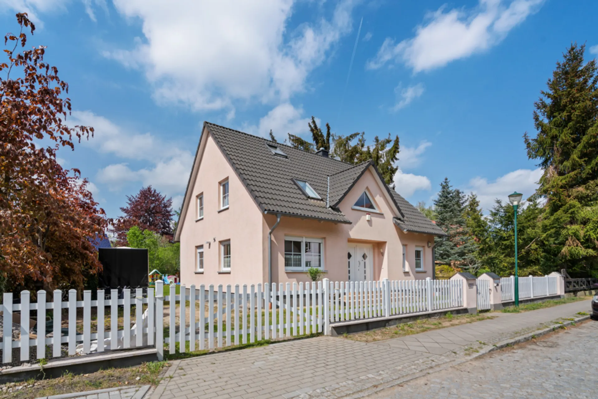 Zweistöckiges Einfamilienhaus mit hellrosa Fassade, grauem Dach und weißem Zaun, umgeben von Bäumen.