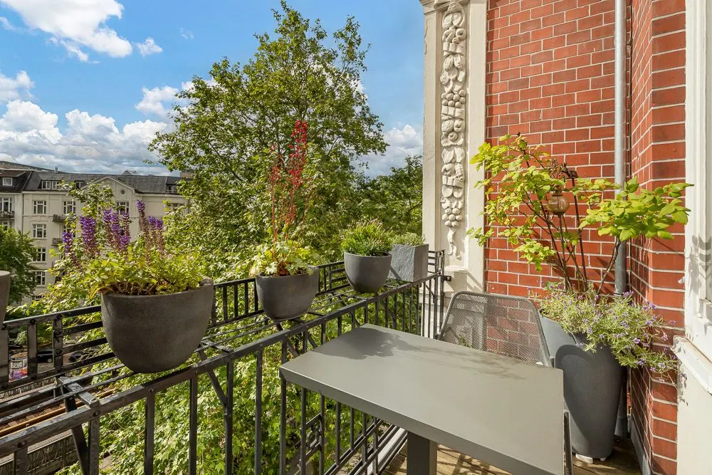 Kleiner Balkon mit Topfpflanzen, Tisch und Stuhl vor einer roten Backsteinfassade.