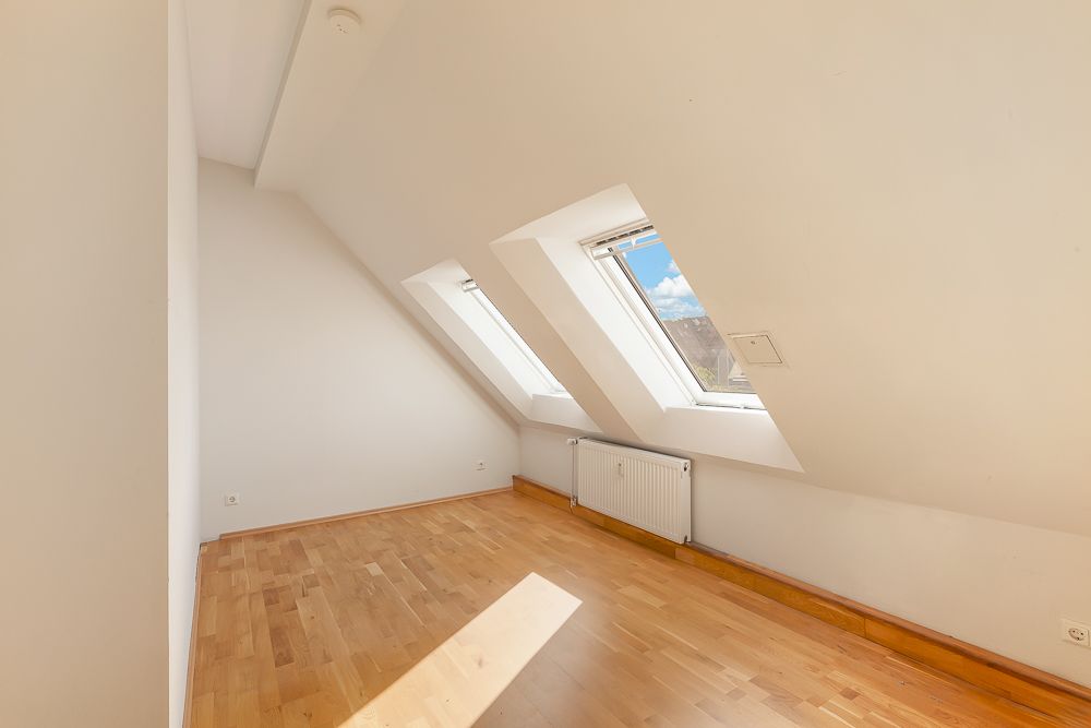 Helles Dachzimmer mit Holzfußboden, zwei Dachfenstern und Heizkörper unter den Fenstern.