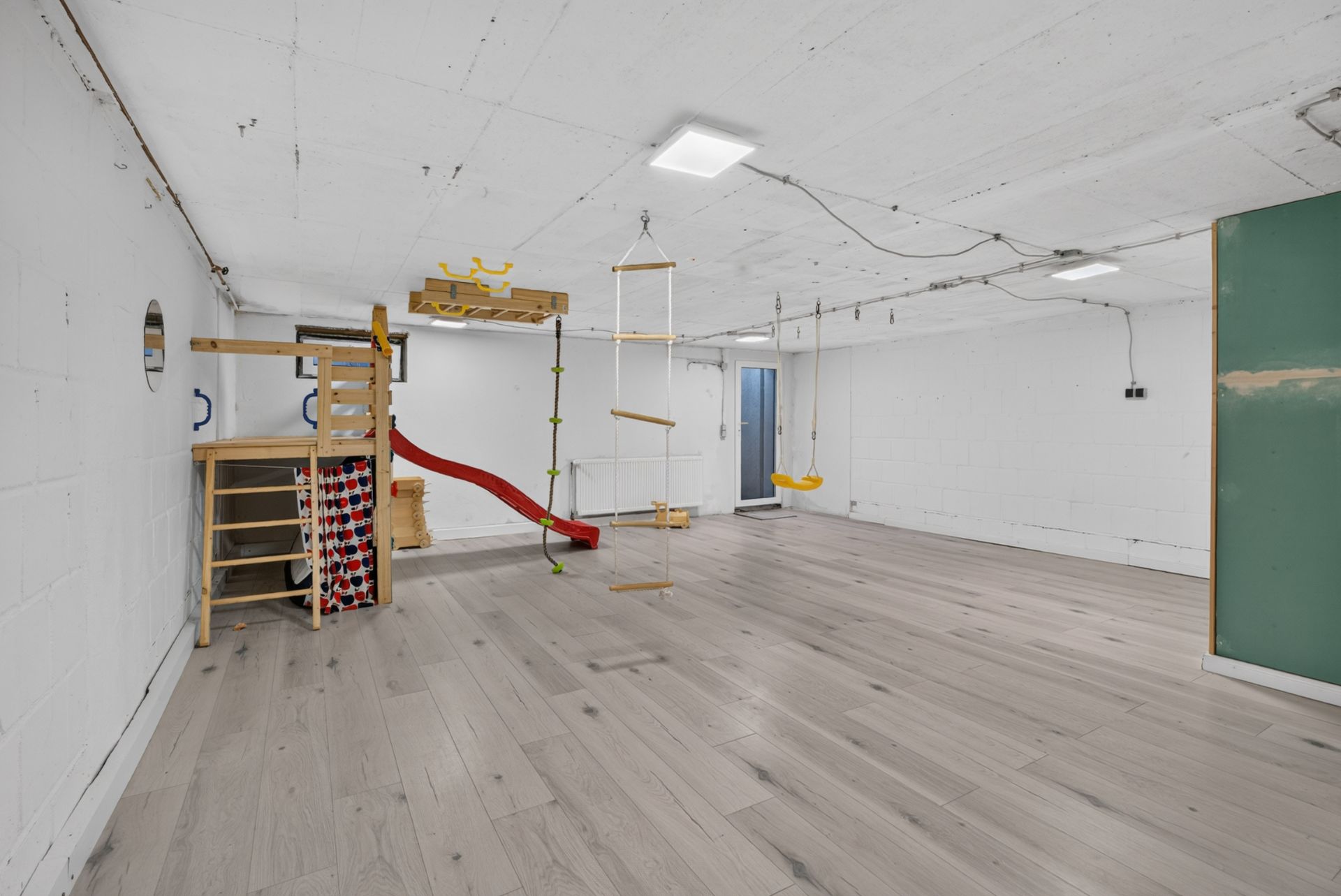 Heller Raum mit Spielgerüst, Rutsche, Schaukel und Holzleiter auf hellem Holzboden.