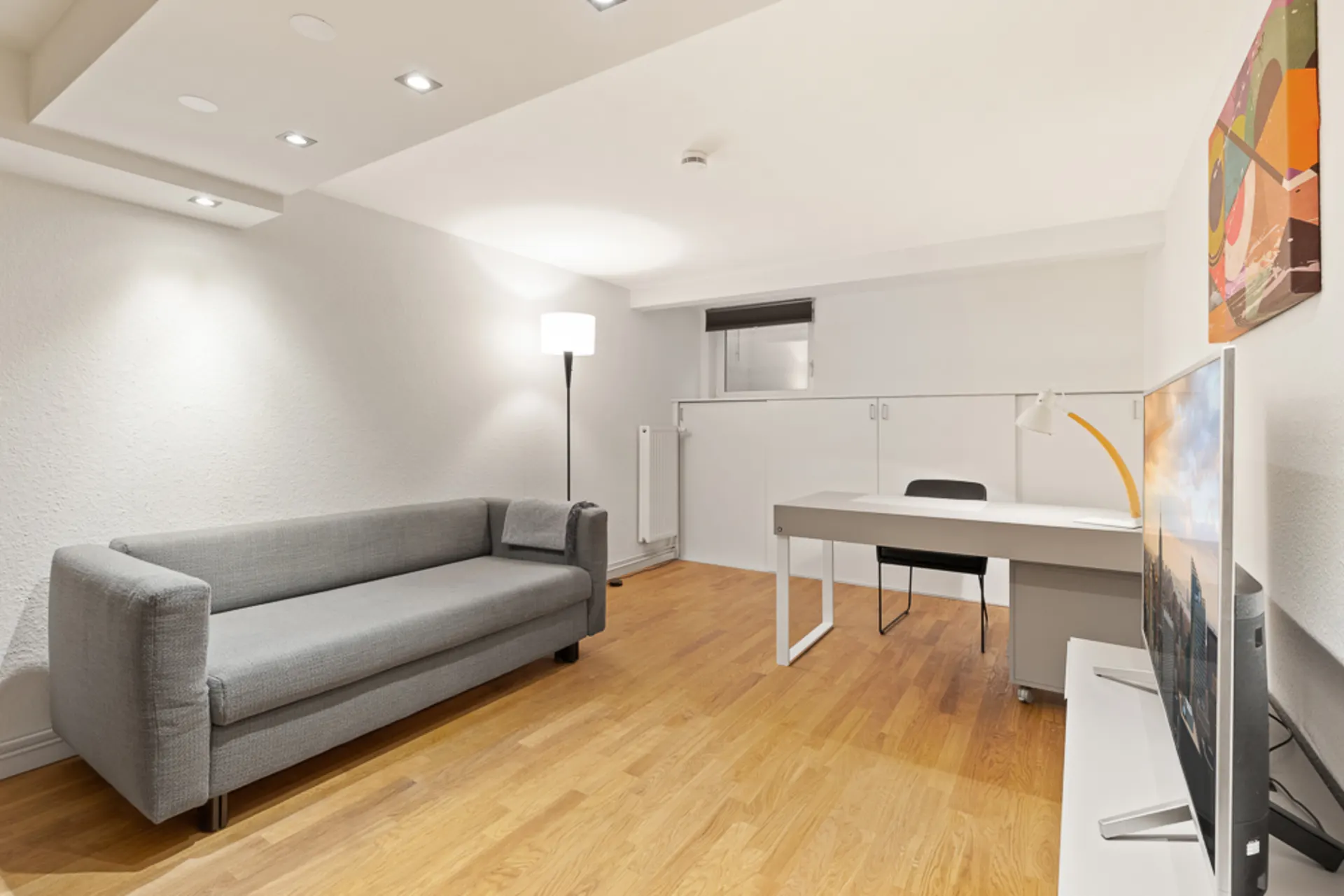 Modernes Zimmer mit grauem Sofa, Schreibtisch, Fernseher und Stehlampe auf Holzfußboden.