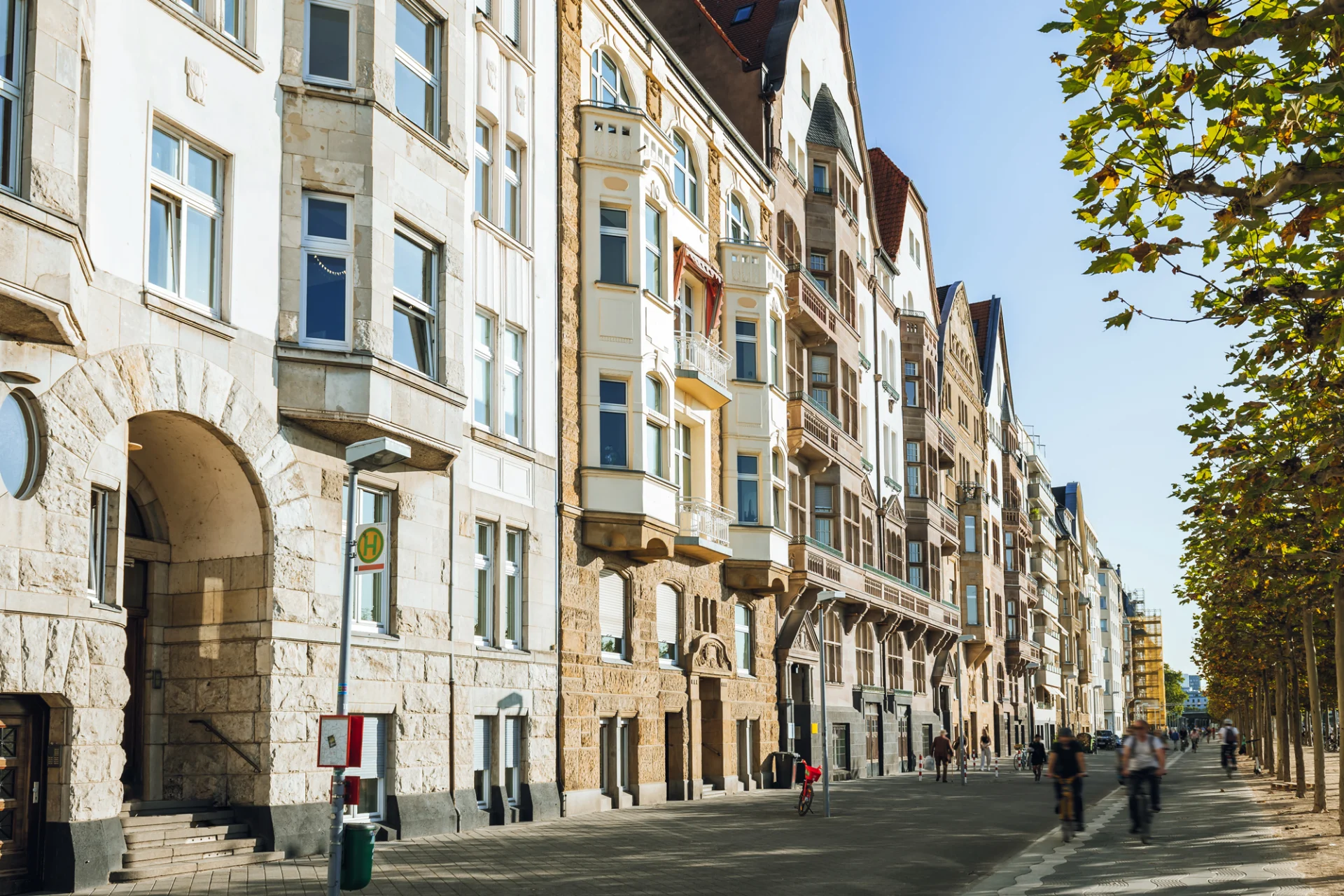 Mehrstöckige Wohnhäuser mit Balkonen an einer von Bäumen gesäumten Straße bei Sonnenschein.