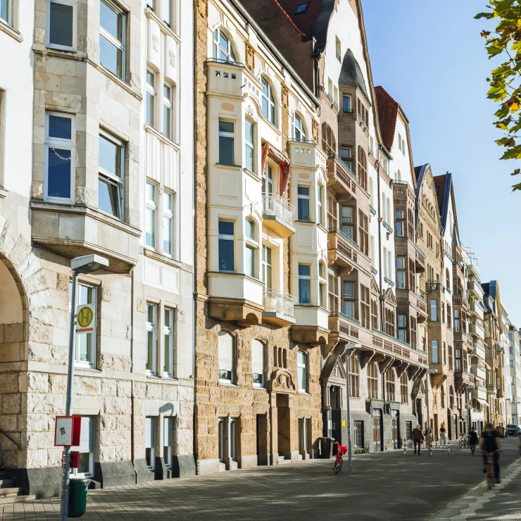 Mehrstöckige Wohnhäuser mit Balkonen an einer von Bäumen gesäumten Straße bei Sonnenschein.