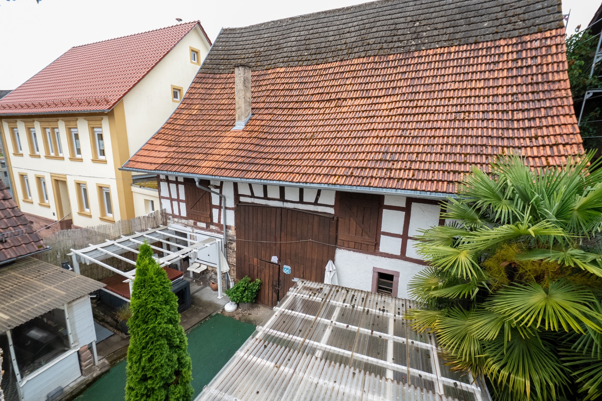 Innenhof mit Fachwerkhaus, roten Dachziegeln, überdachtem Bereich und Palmenpflanze.