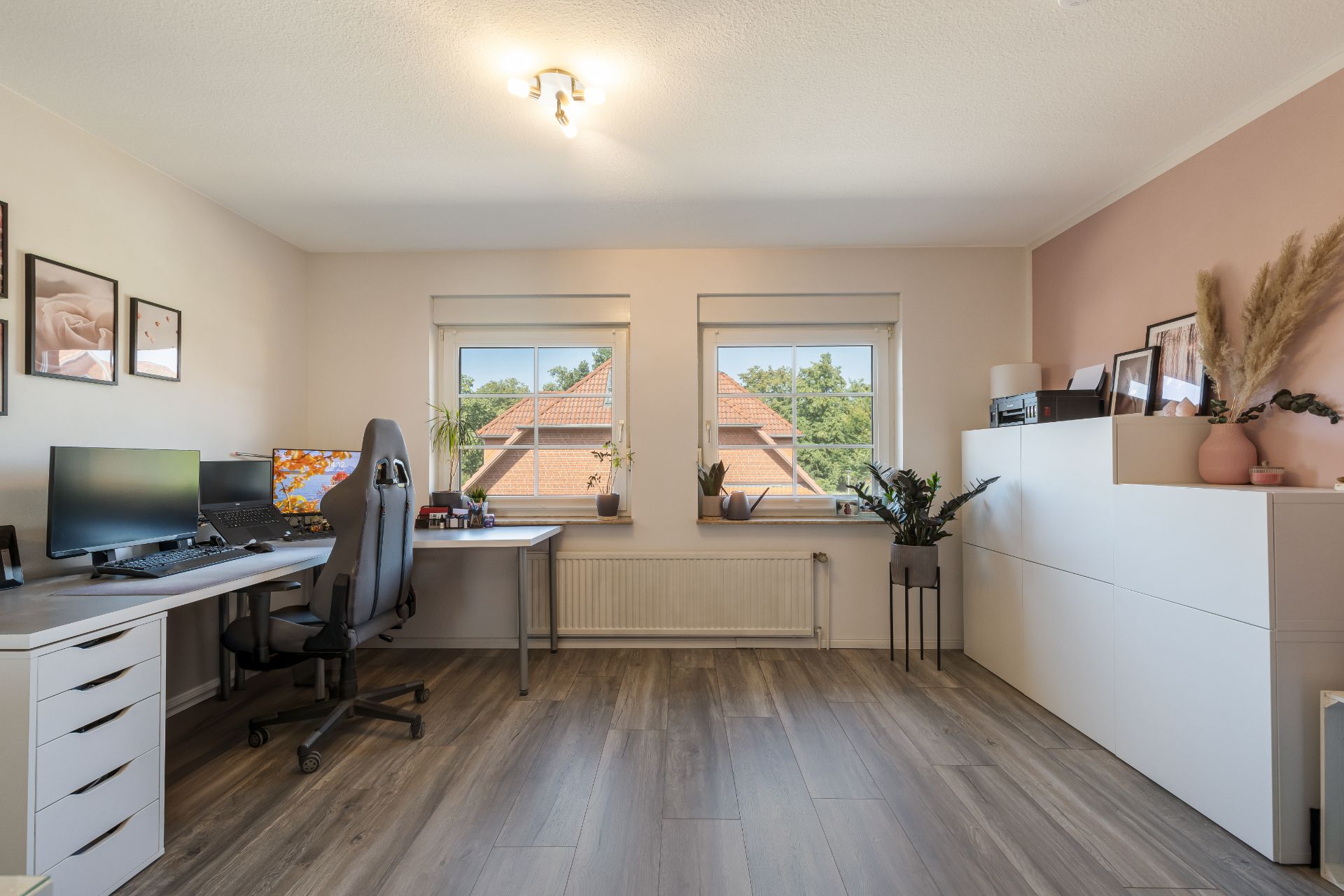 Helles Arbeitszimmer mit zwei Schreibtischen, Stuhl, Pflanzen und großen Fenstern.