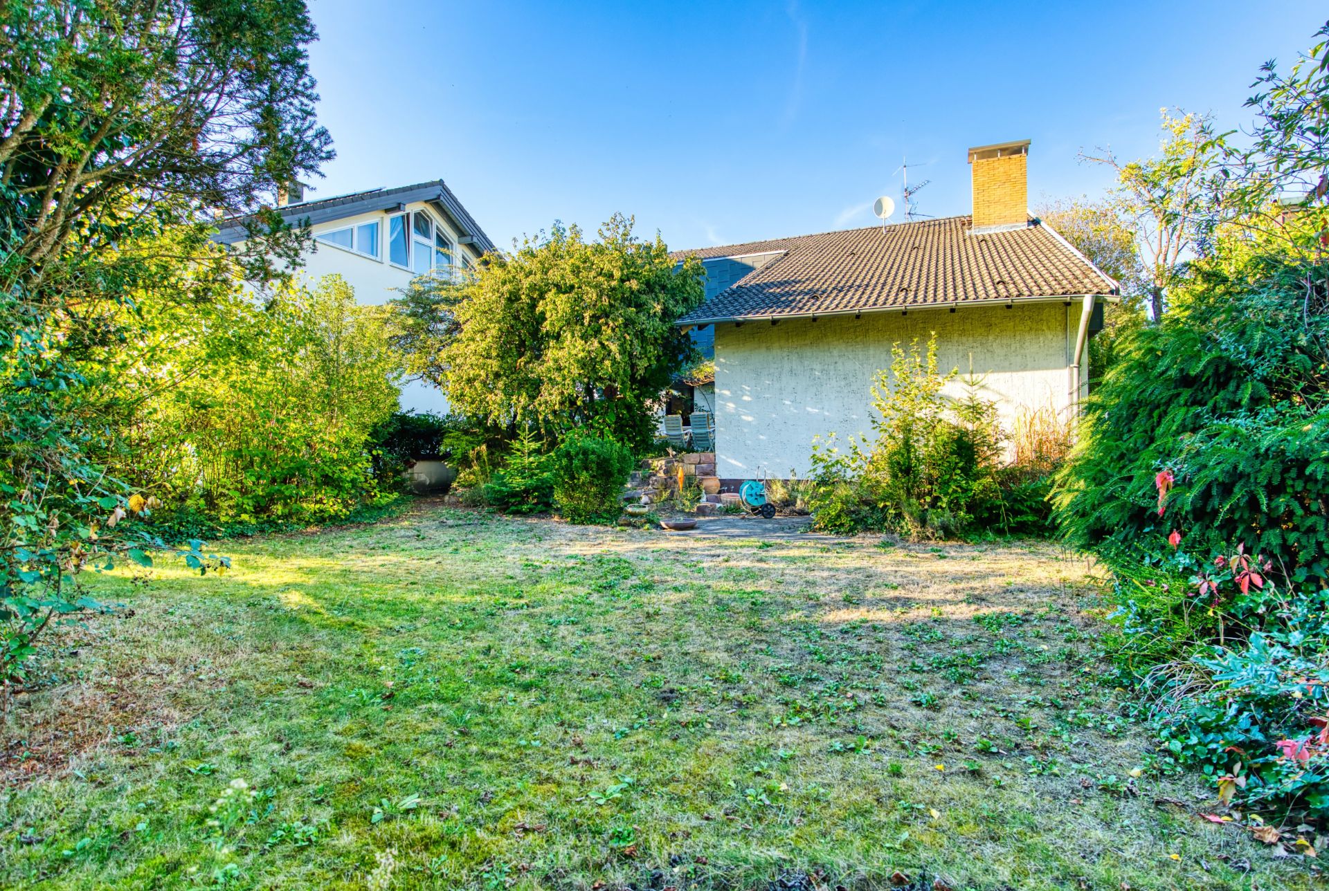 Garten mit Rasenfläche, Bäumen und Sträuchern hinter einem Wohnhaus bei Tageslicht.