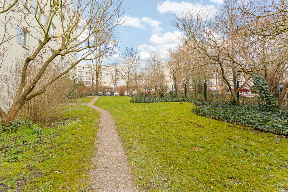 Grüner Gartenbereich mit Gehweg, Bäumen und einigen parkenden Autos im Hintergrund.