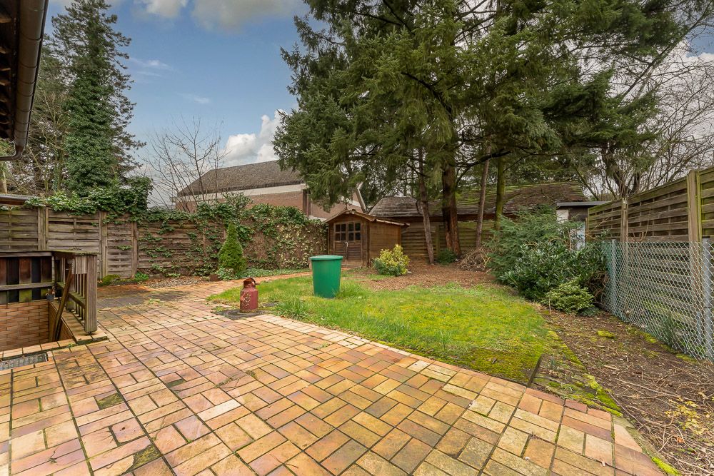 Garten mit Terrasse, Rasen, Bäumen, Holzzaun und kleinem Holzhaus im Hintergrund.