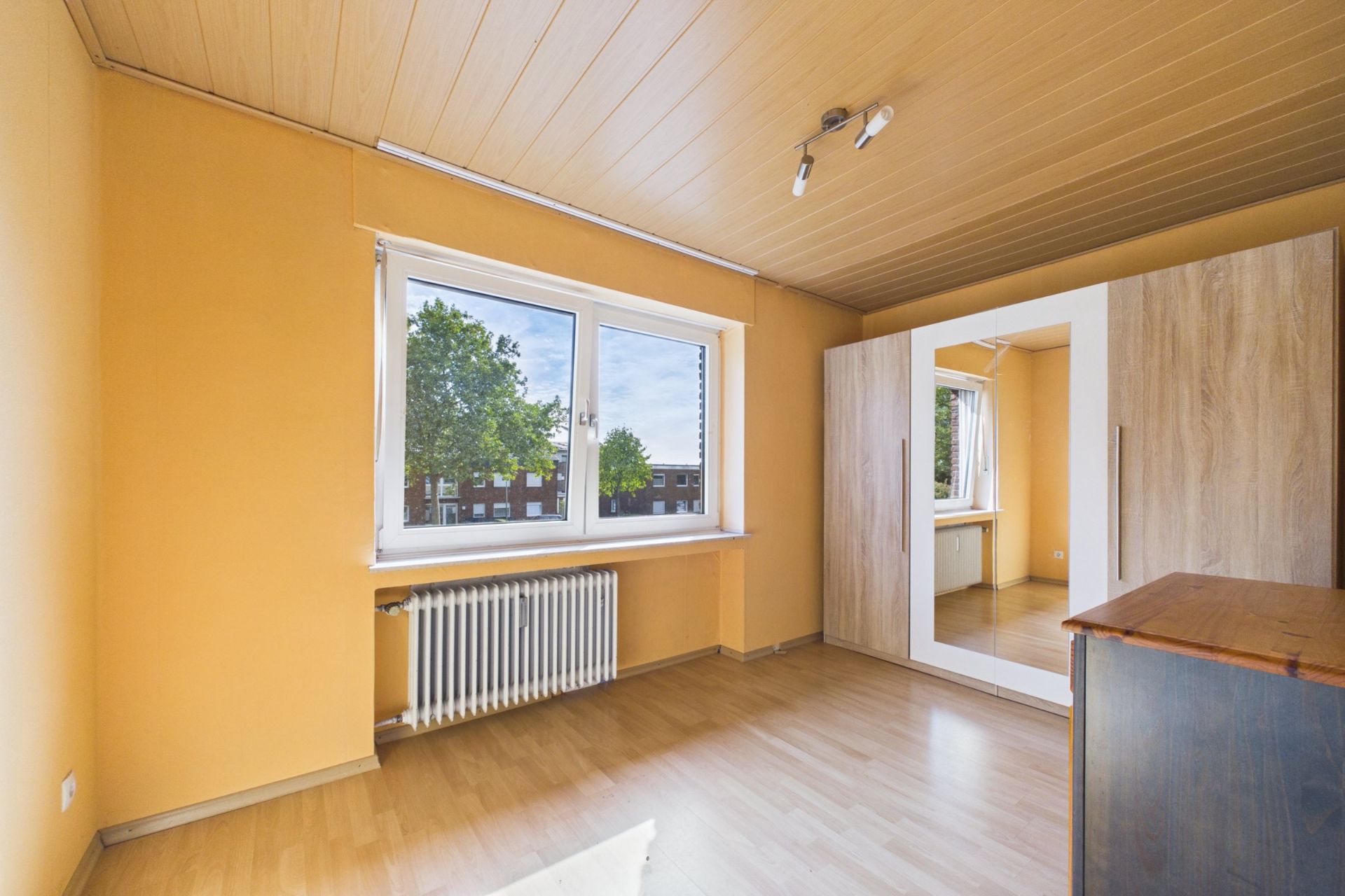 Leeres Zimmer mit Holzdecke, großem Fenster, Heizkörper, Kleiderschrank und Kommode.